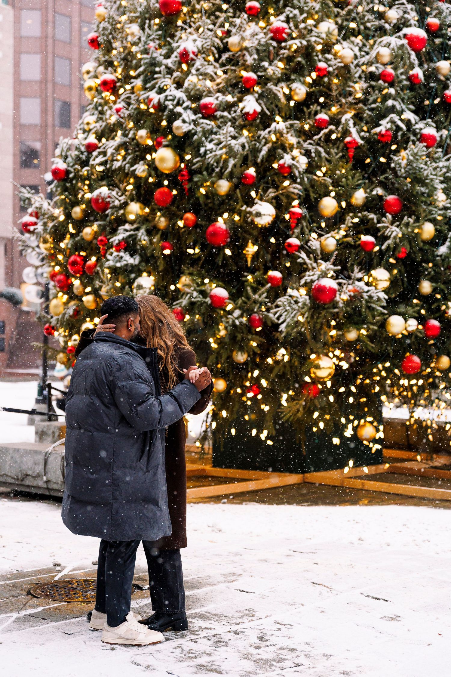 Couple standing in front of a large decorated Christmas tree with red and gold ornaments in a snowy outdoor setting.