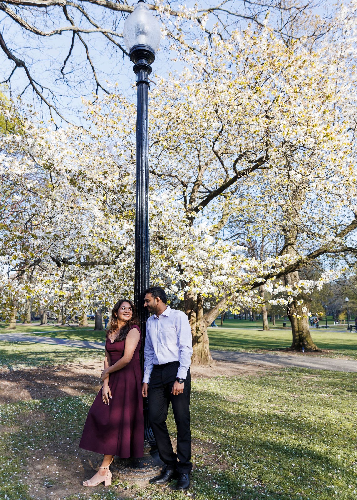 Couple leaning against a lamp post in a park with blooming white trees, woman in burgundy dress, man in white shirt.