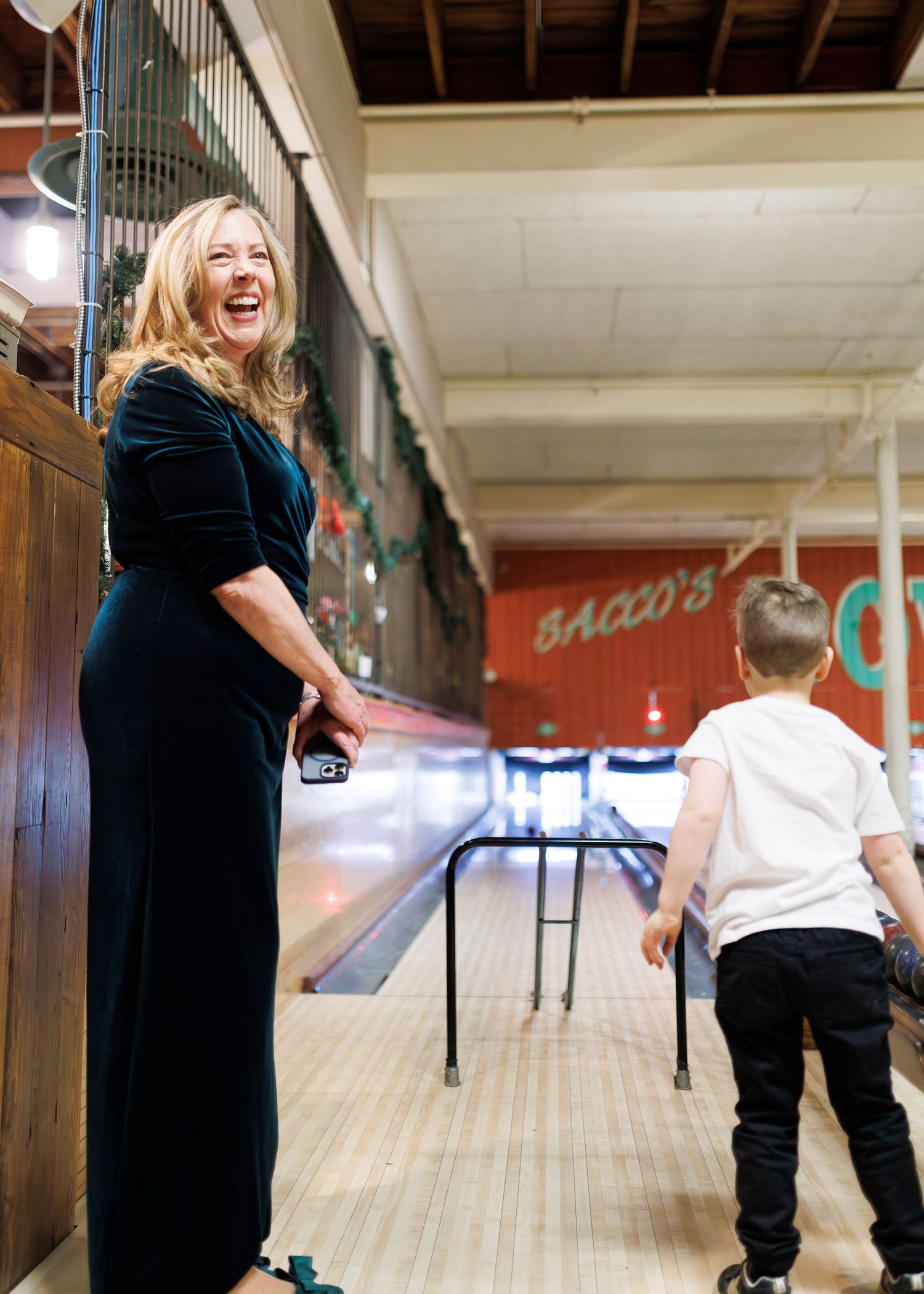 A joyful moment at a bowling alley shows a person in a dark velvet dress laughing while a child stands nearby at Flatbreads in Somerville.