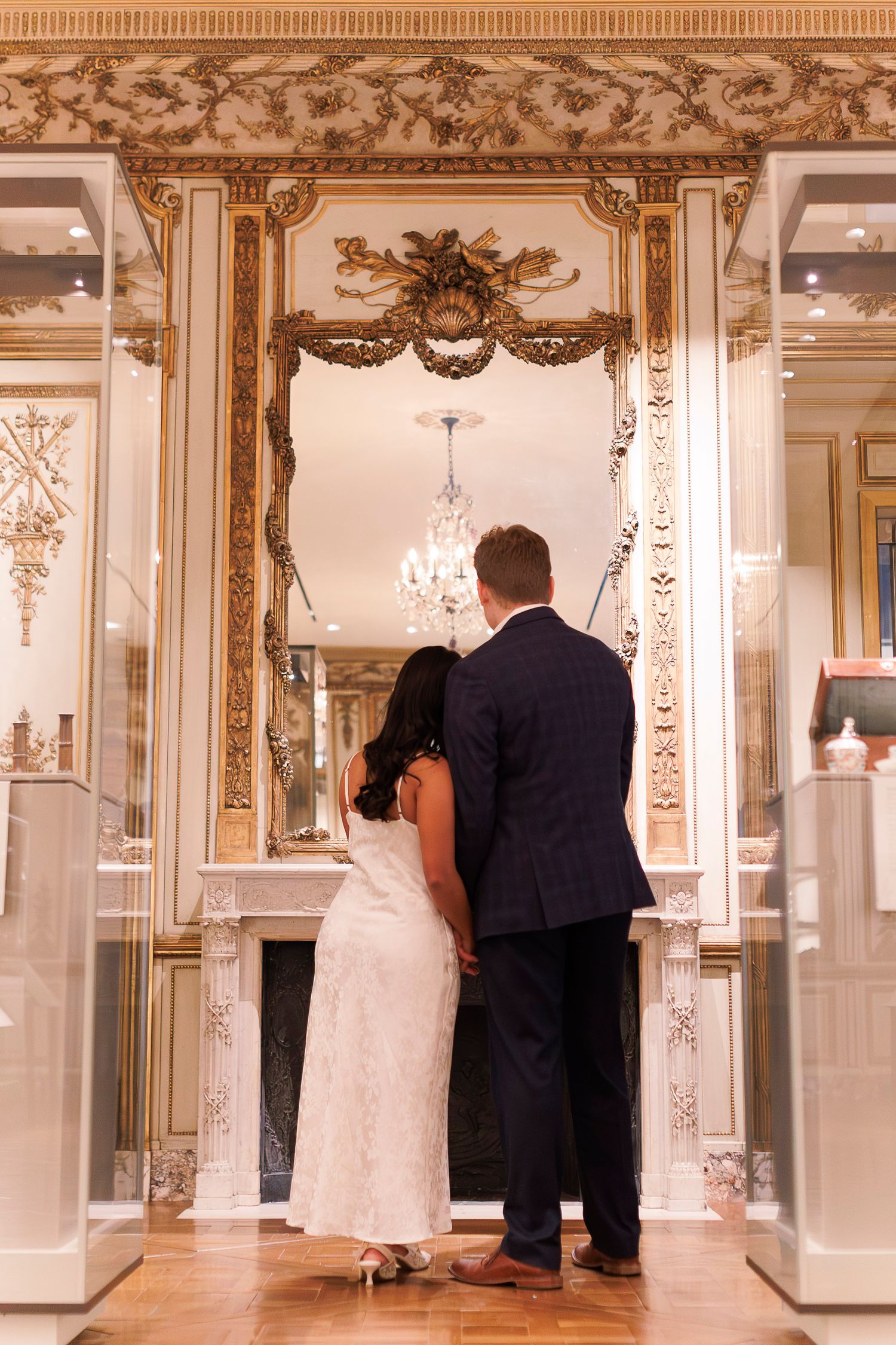 A couple stands in an elegant ballroom with ornate gold trim and mirrors during a formal event.