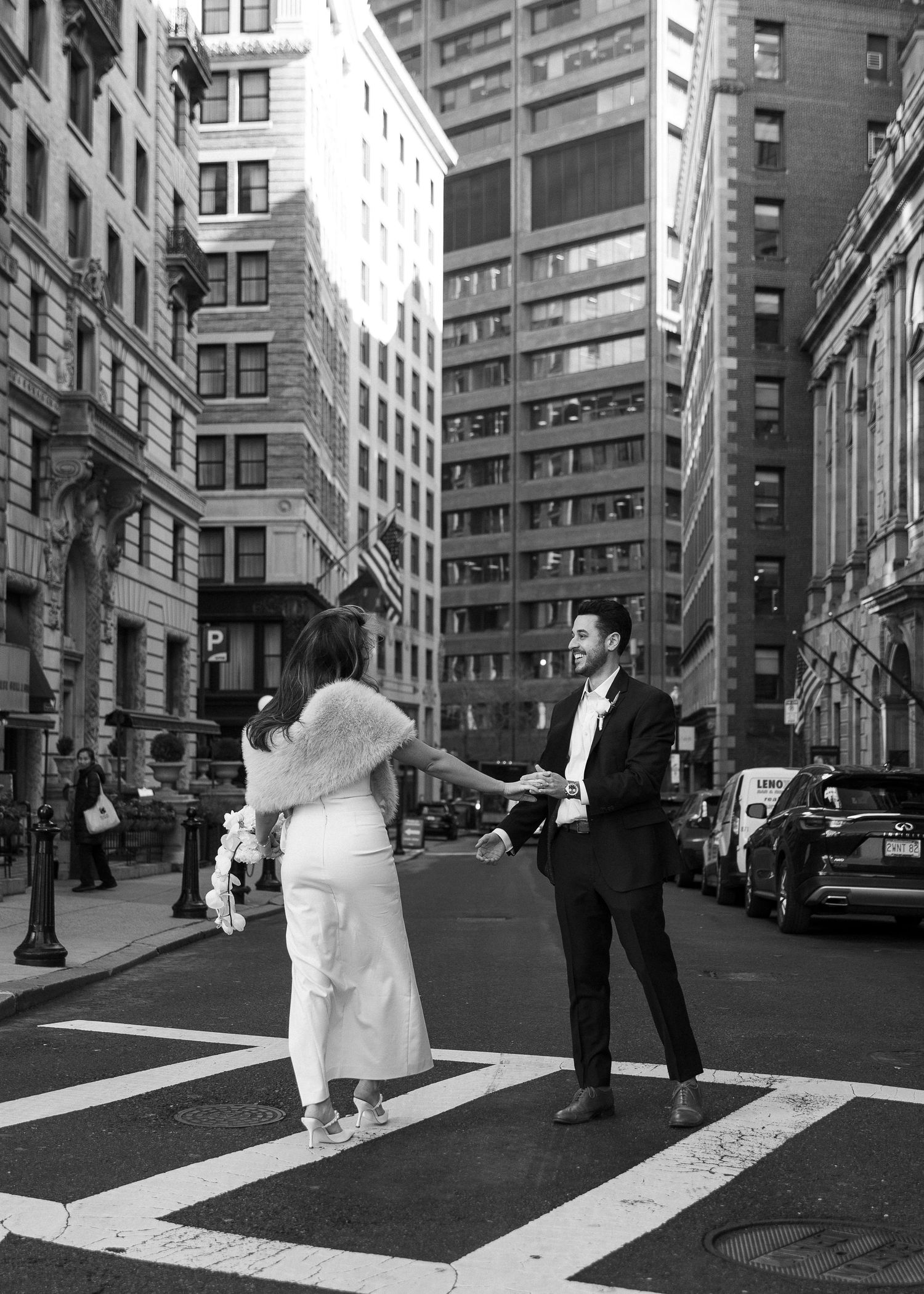 Black and white photo of a couple in wedding attire holding hands on a crosswalk in New York City.