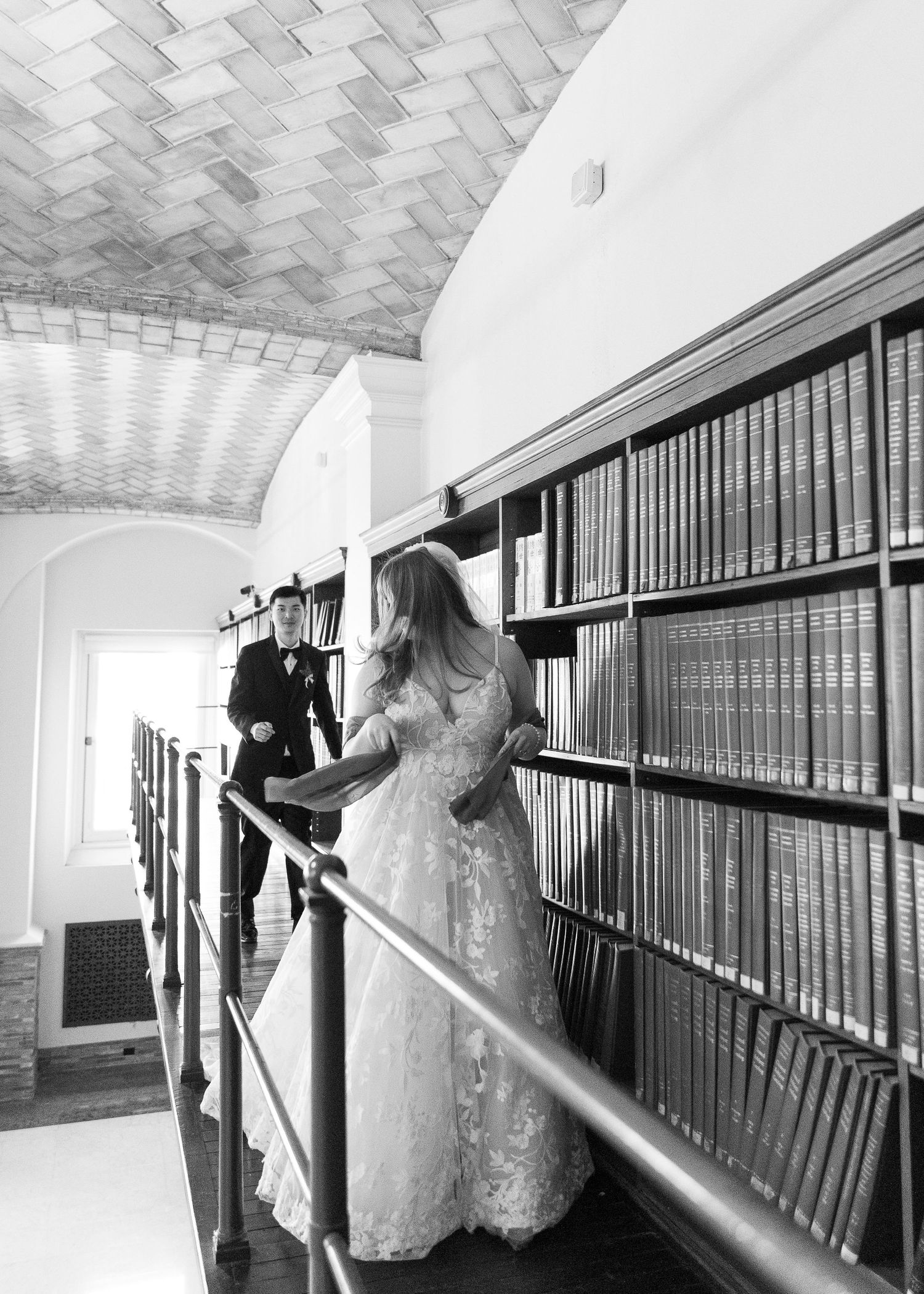 Bride in lace gown and groom in tuxedo walk along a grand library bookshelf in black and white wedding photo.