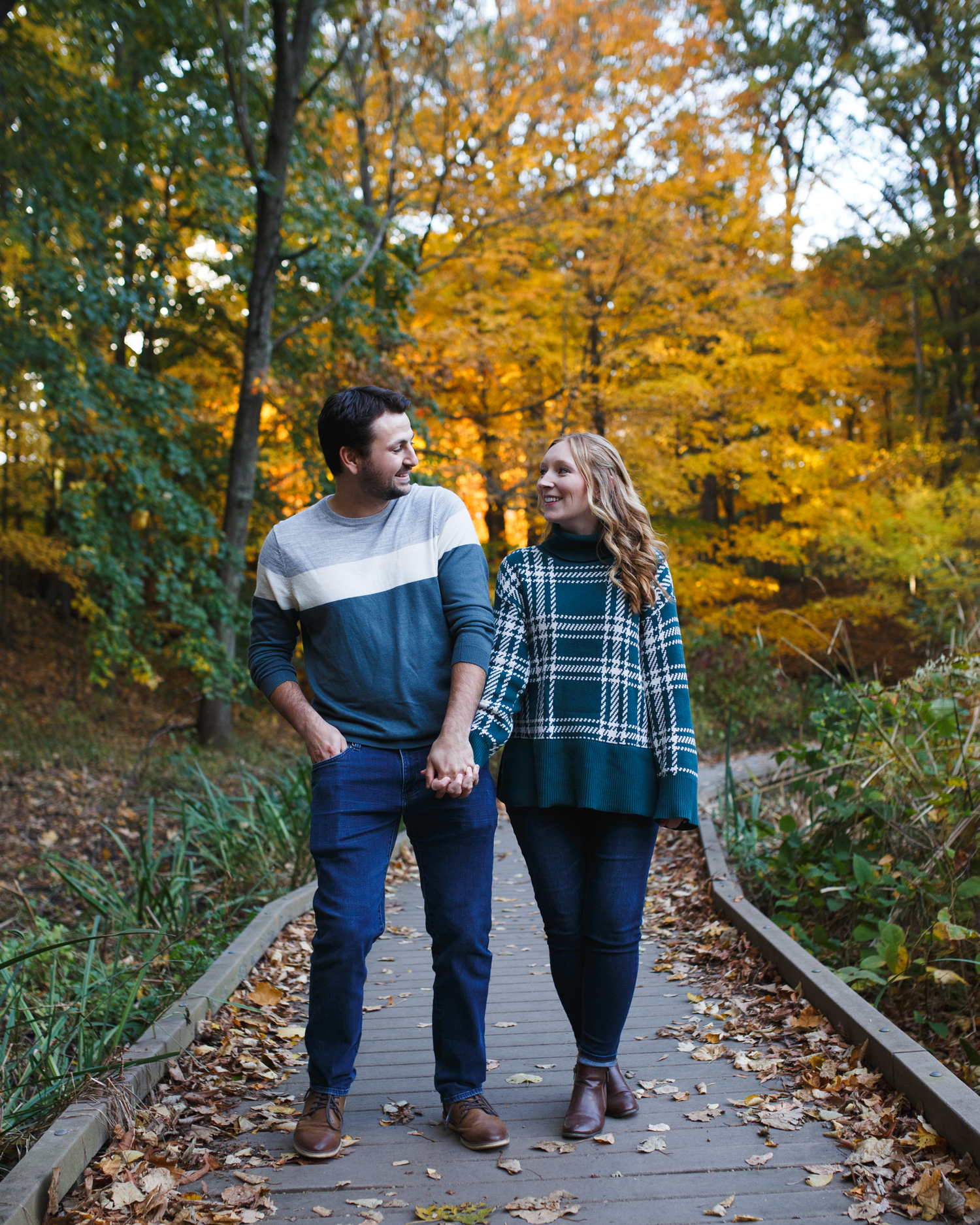 Couple holding hands walking on a wooden boardwalk surrounded by vibrant autumn foliage in fall colors.