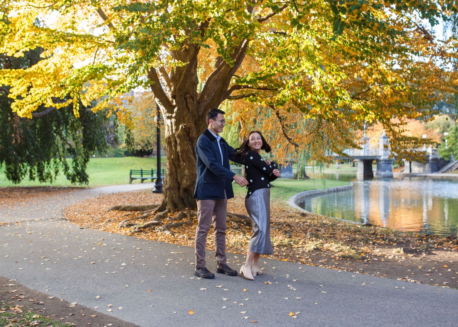 A couple walks their dog along a tree-lined path with golden autumn foliage overhead on a sunny day.
