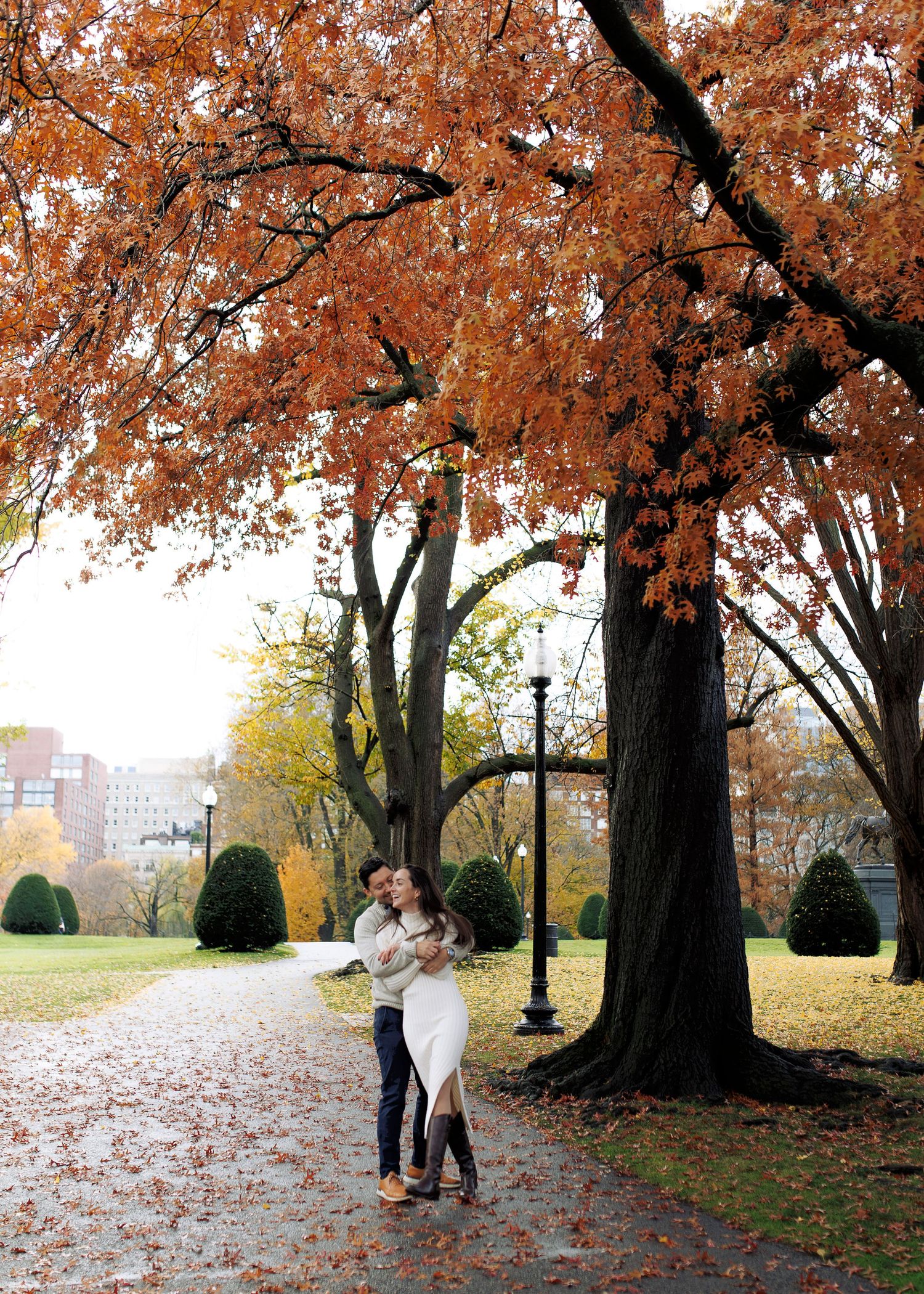 Two figures embrace under autumn trees with fallen leaves scattered on the ground in a park setting.