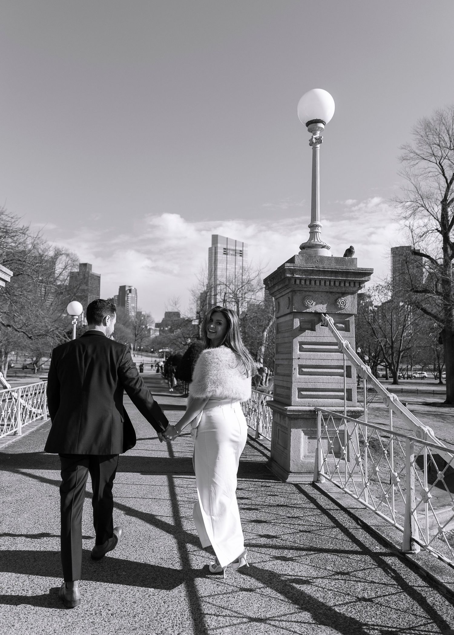 A black and white photo series showing a couple walking along a park path with city buildings visible in the background.