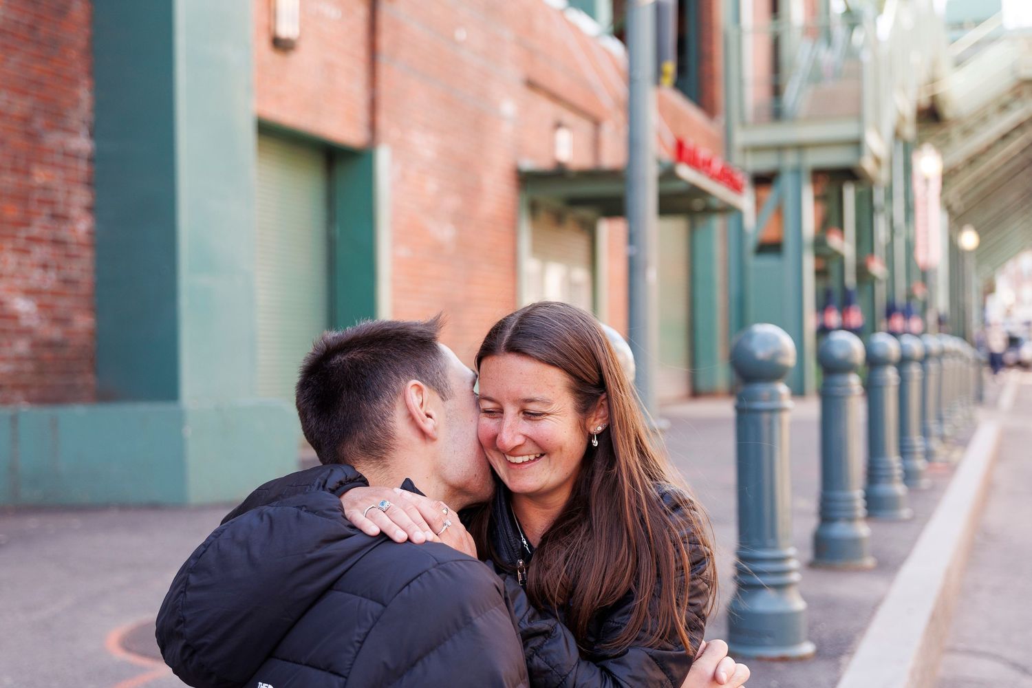 Couple sharing a romantic moment outdoors, woman smiling joyfully as man kisses her cheek near brick buildings.