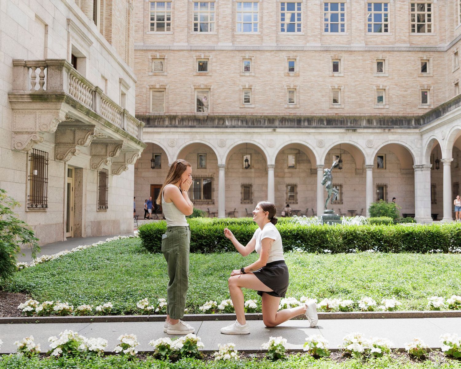 A person proposes on one knee to their partner in a sunlit courtyard garden of a classical stone building.