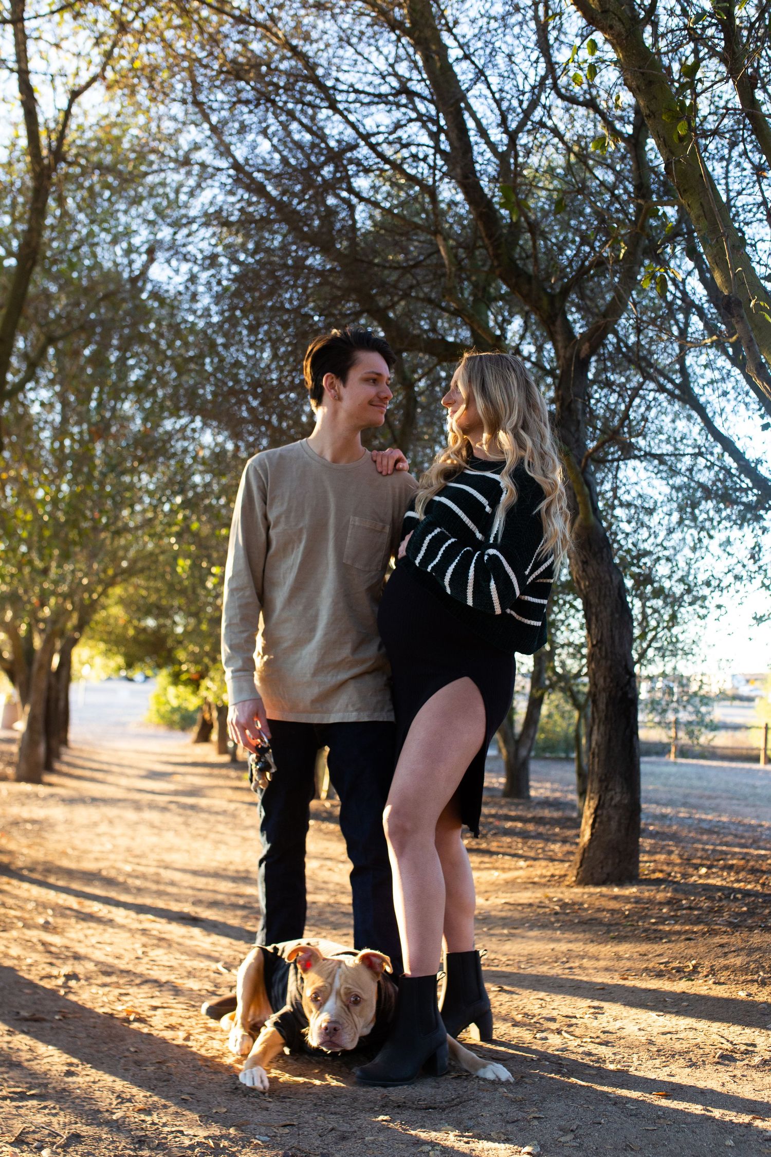 Couple playfully interacting in a sunlit outdoor setting with trees casting long shadows on the ground.