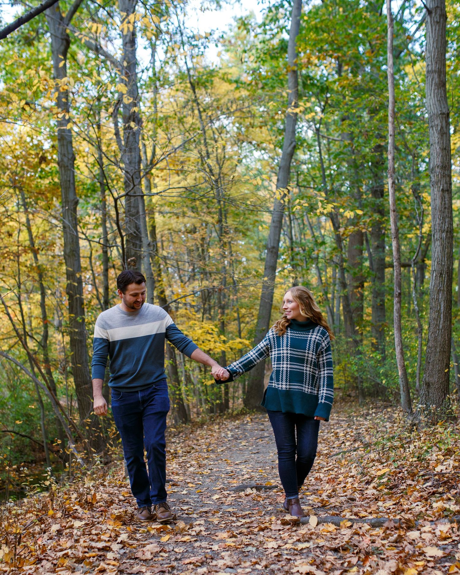 A couple walks along a leaf-covered forest path during autumn with golden foliage overhead.