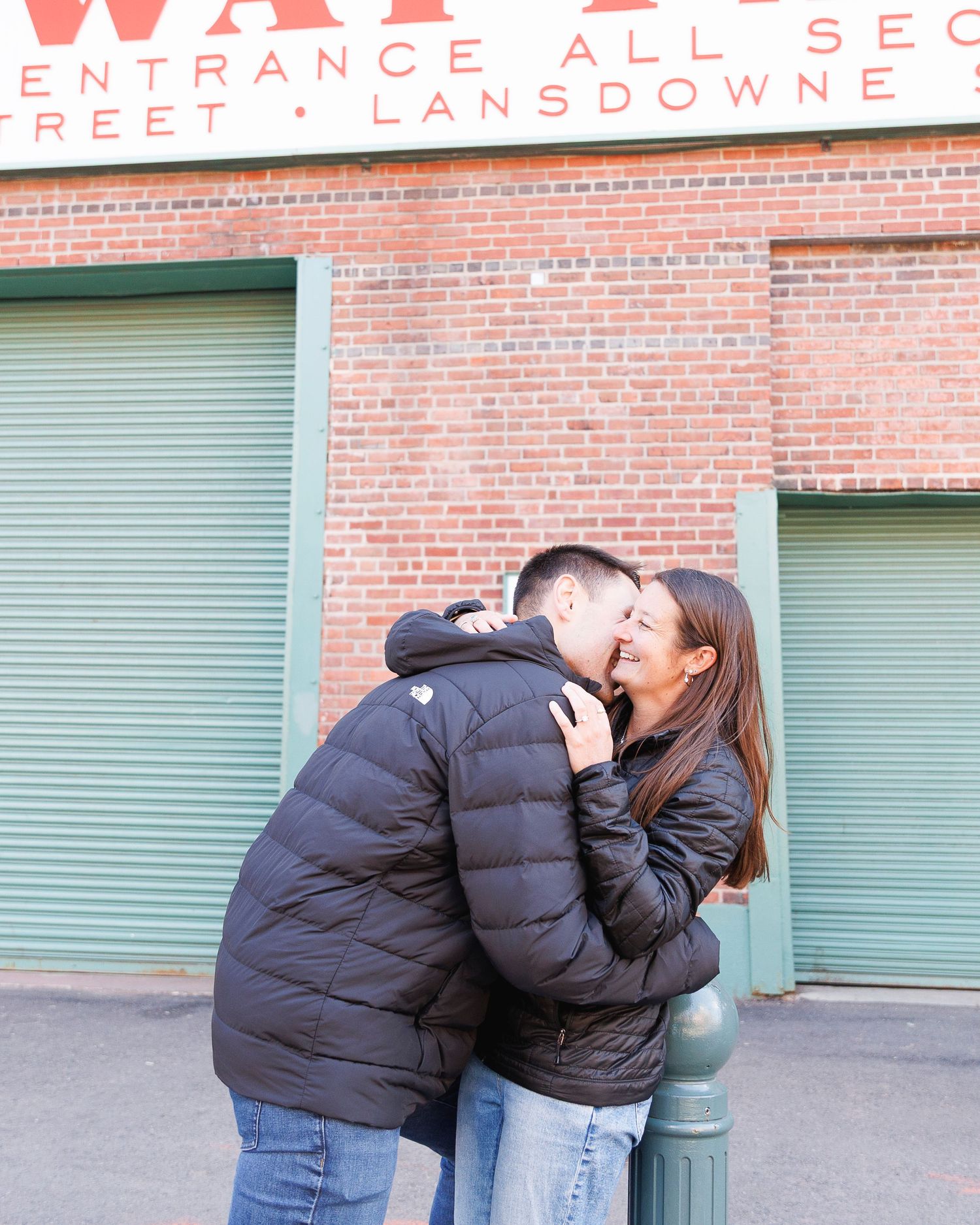 Couple sharing a romantic kiss outside Fenway Park on Lansdowne Street, both wearing black puffer jackets.