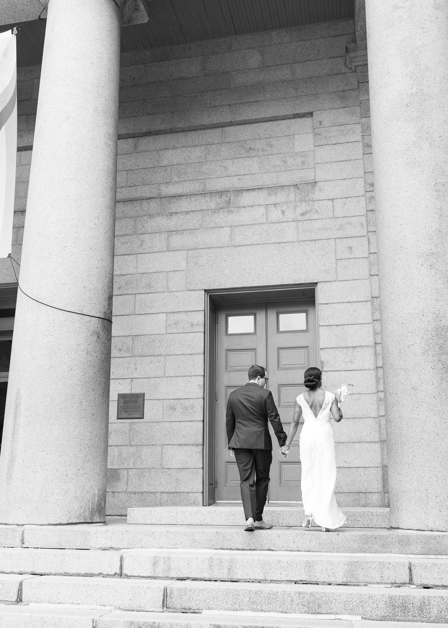 Black and white photo of a bride and groom holding hands walking up steps toward a grand building entrance.