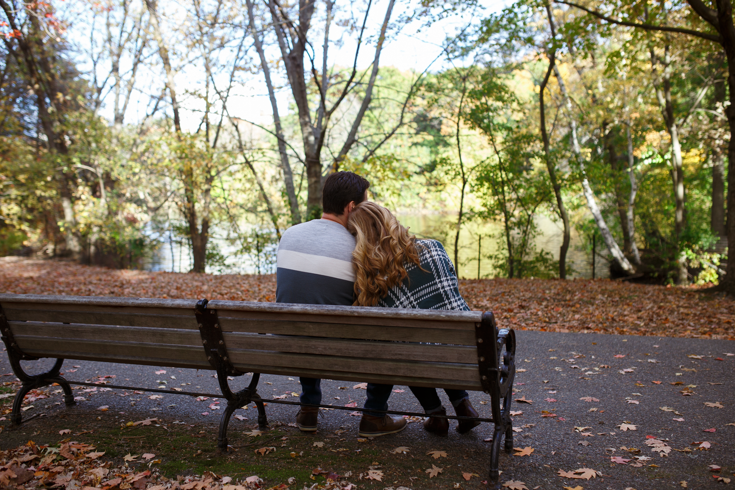 A row of empty wooden benches along a tree-lined path covered in fallen autumn leaves on a sunny day.