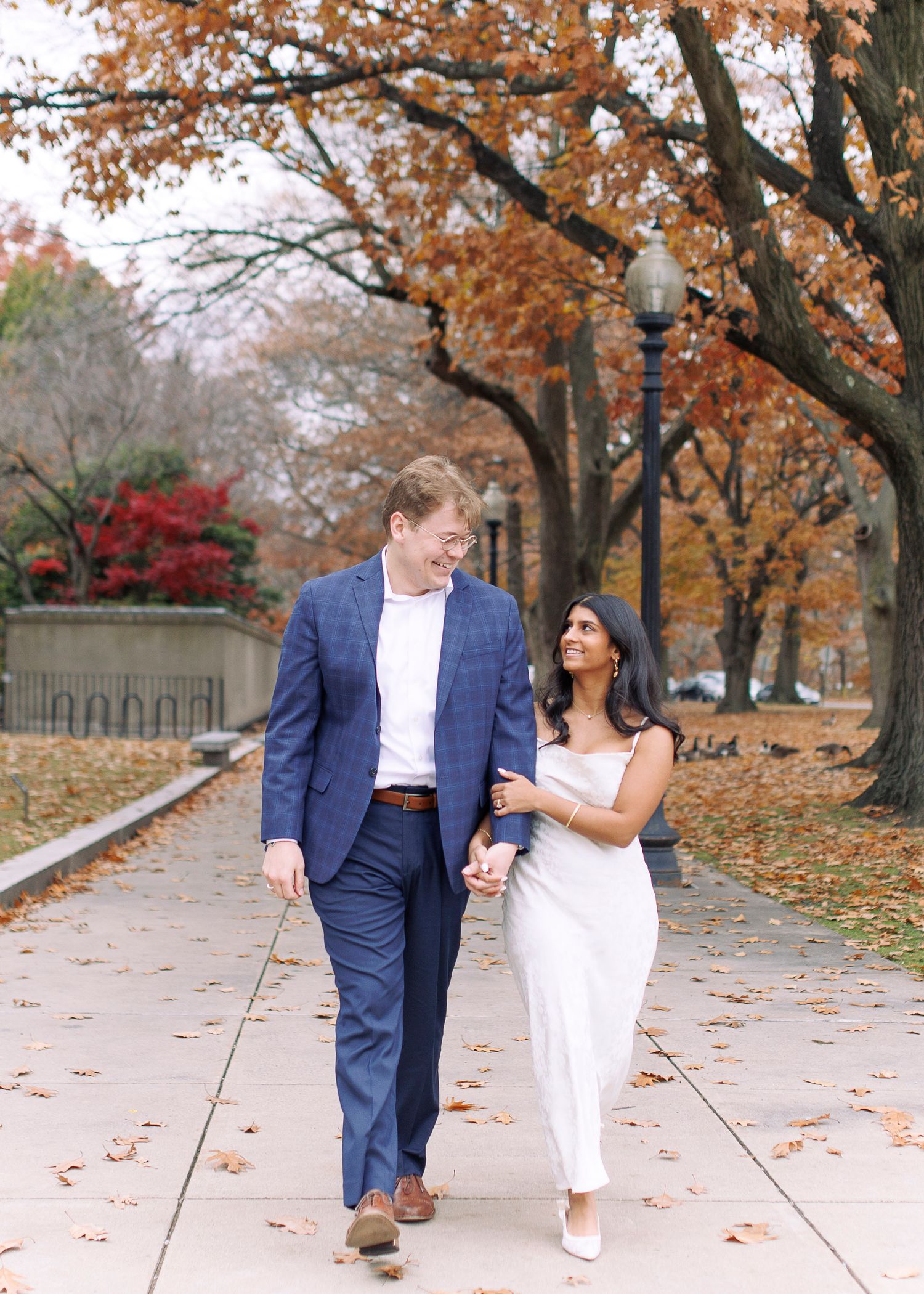 Couple walking together on a tree-lined autumn path with fallen leaves scattered on the ground.