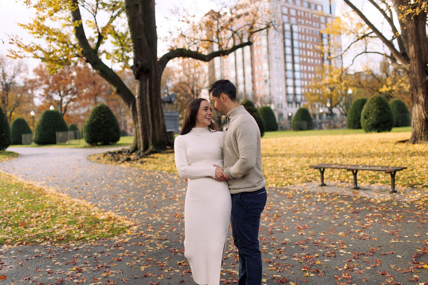 A romantic autumn scene in a park with fall foliage, a winding path, and city buildings visible in the background.