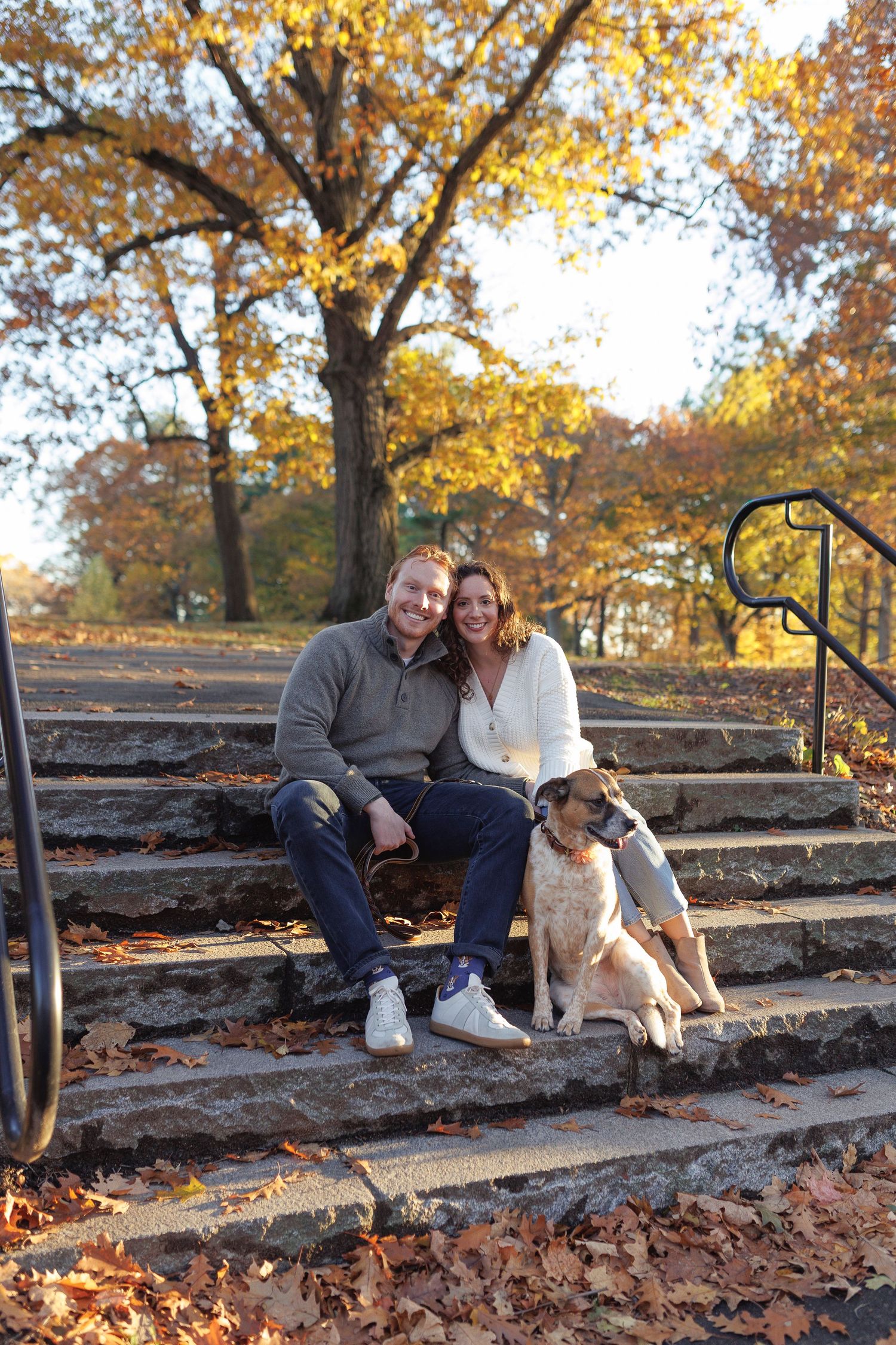 Couple smiling with dog on stone steps surrounded by golden autumn foliage in a park.
