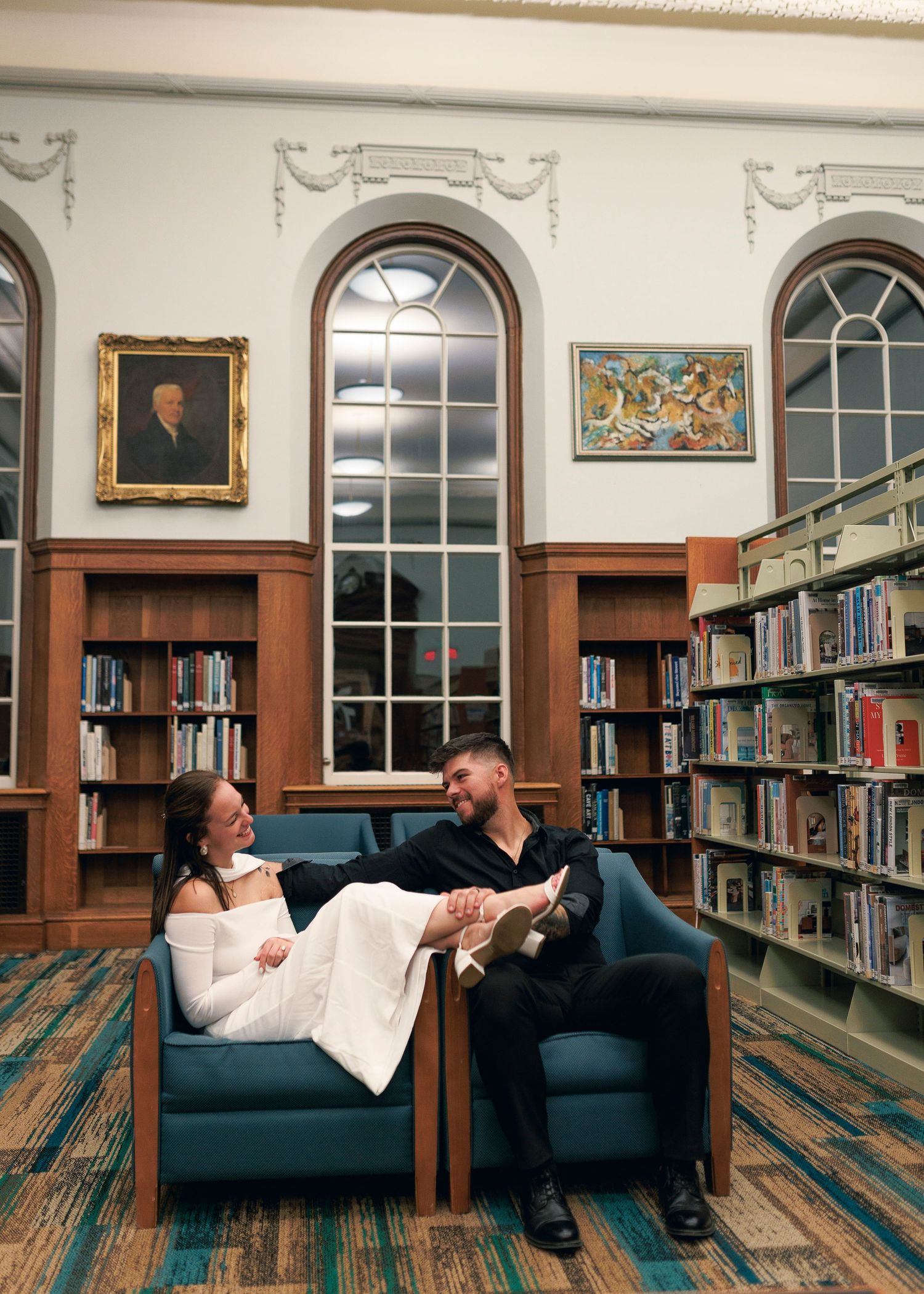 A couple relaxes together on a blue couch in an elegant library with arched windows and wooden bookshelves.