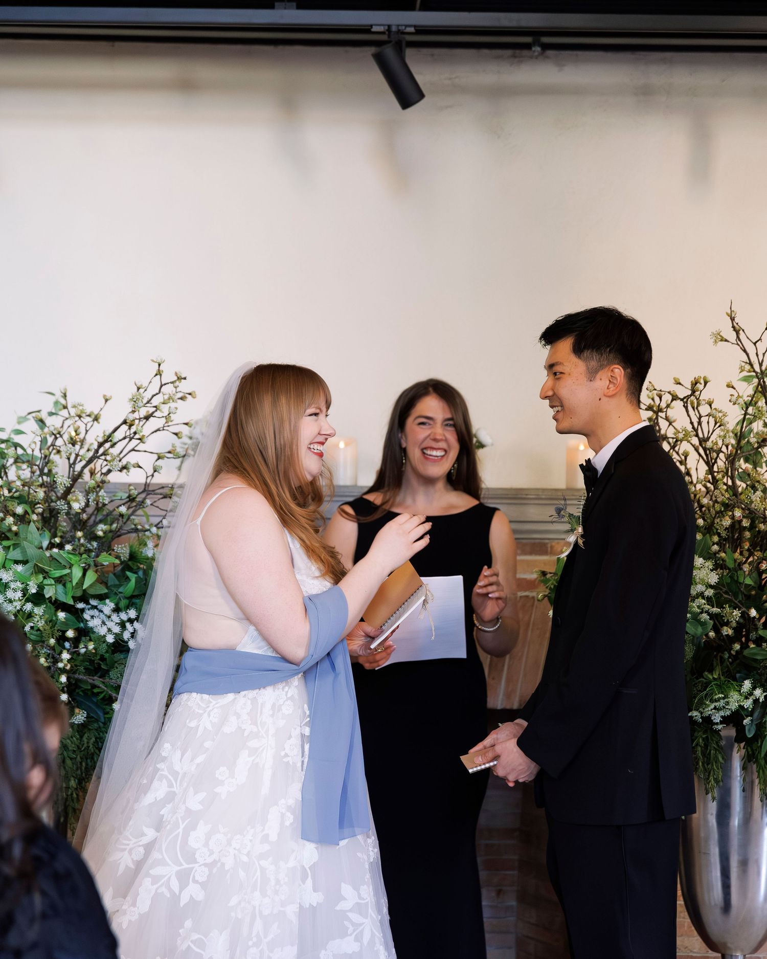 A wedding ceremony takes place in front of a white wall decorated with greenery and florals.