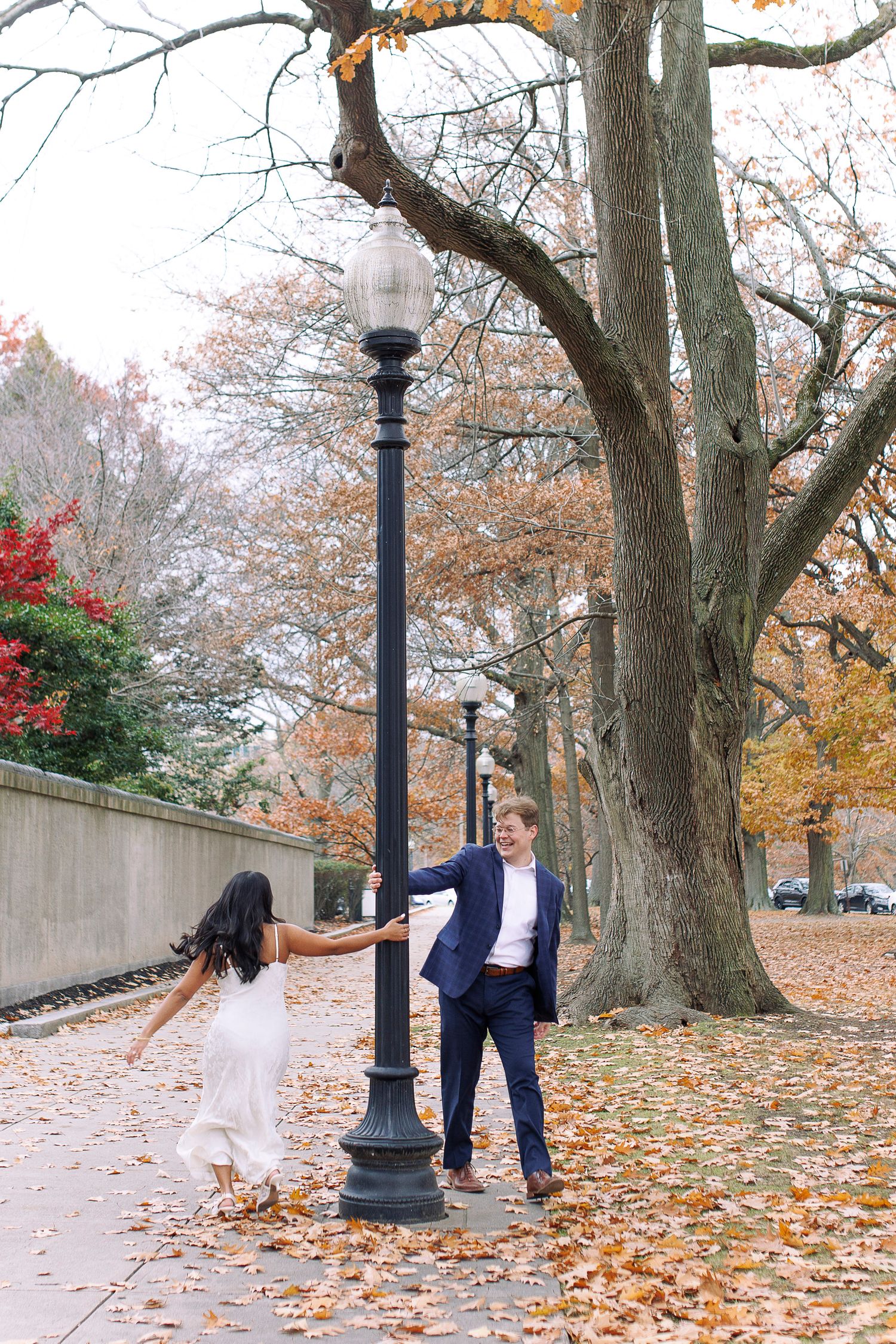 Couple dancing playfully around a lamp post on an autumn day with fallen leaves scattered on the ground.