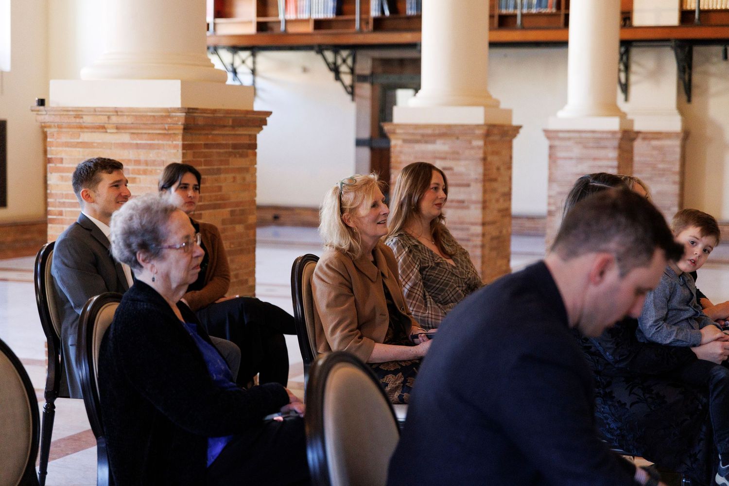 People seated in rows at an indoor event with white columns and brick walls in the background.