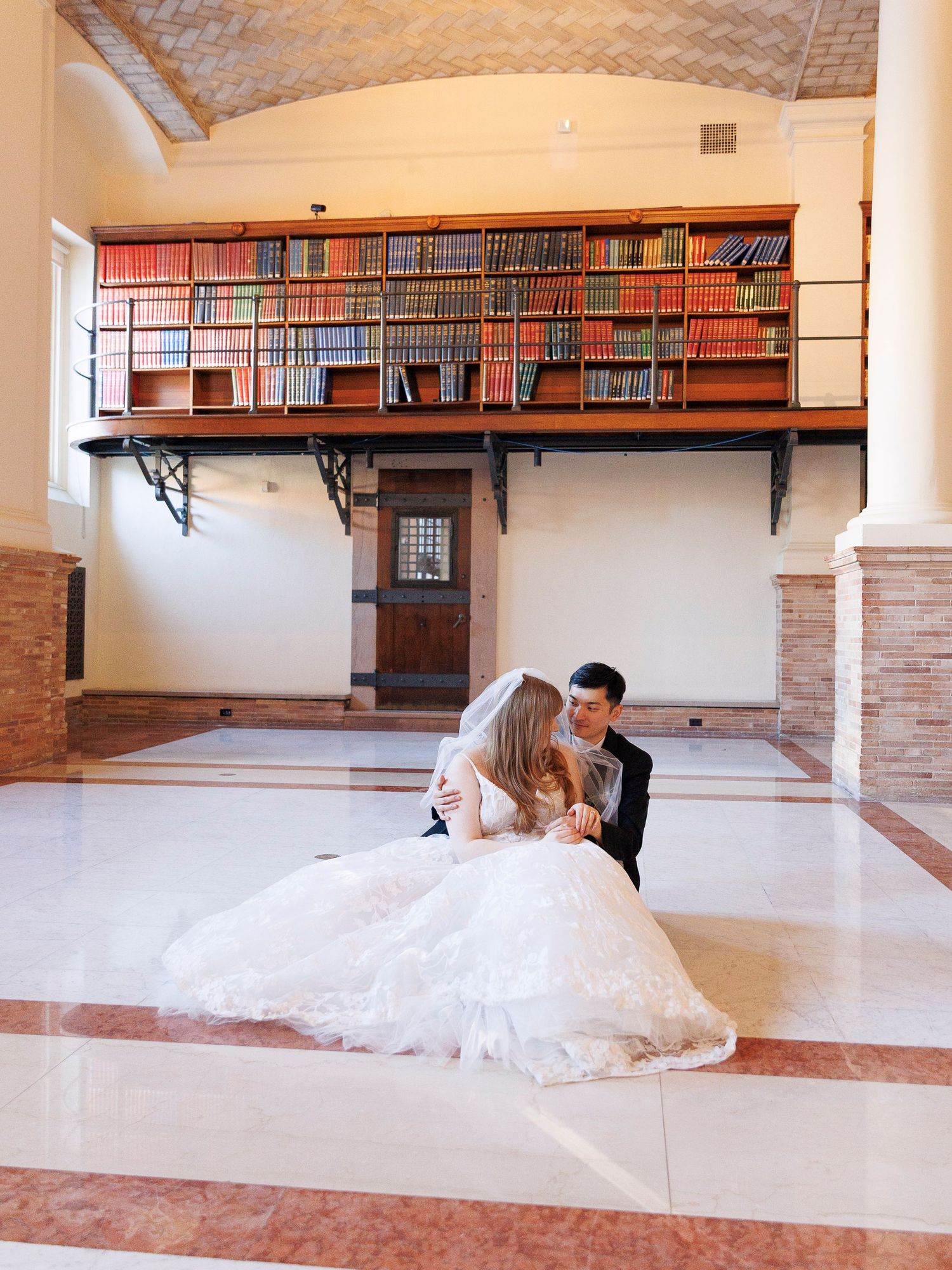 Bride and groom sitting on marble floor in elegant library venue with red bookshelf balcony during wedding portrait session.
