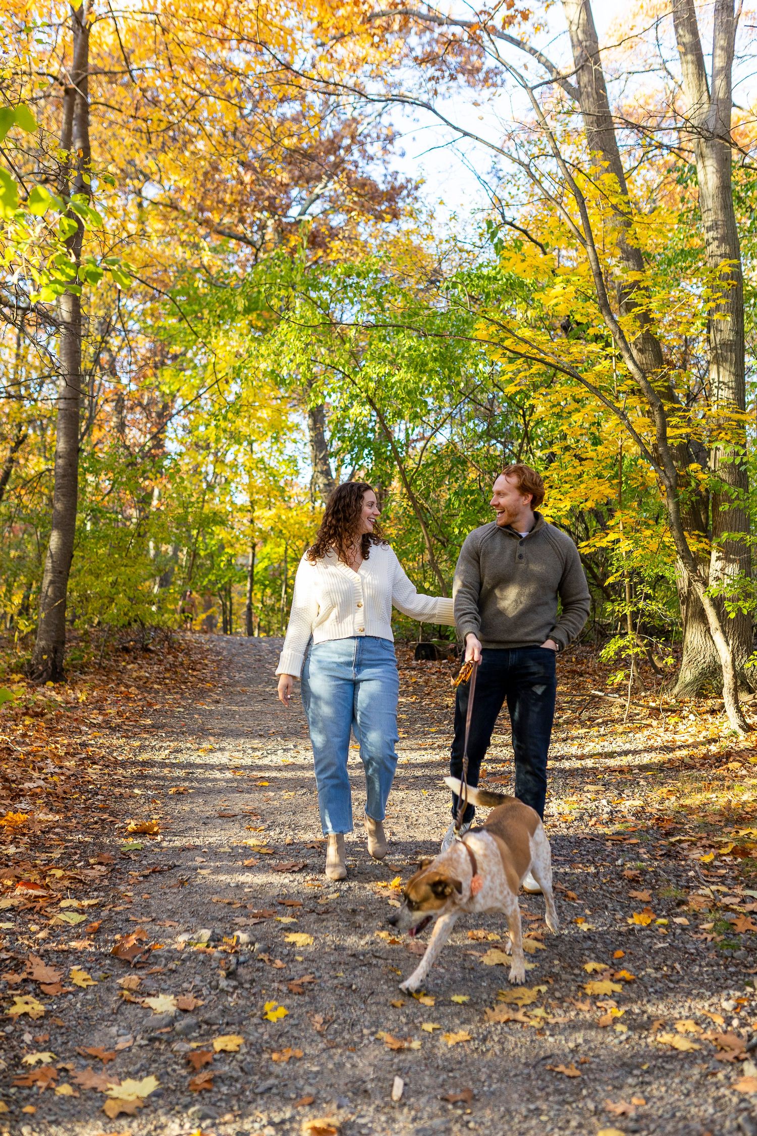 Couple walking a dog on a scenic autumn forest trail surrounded by colorful yellow and green foliage.