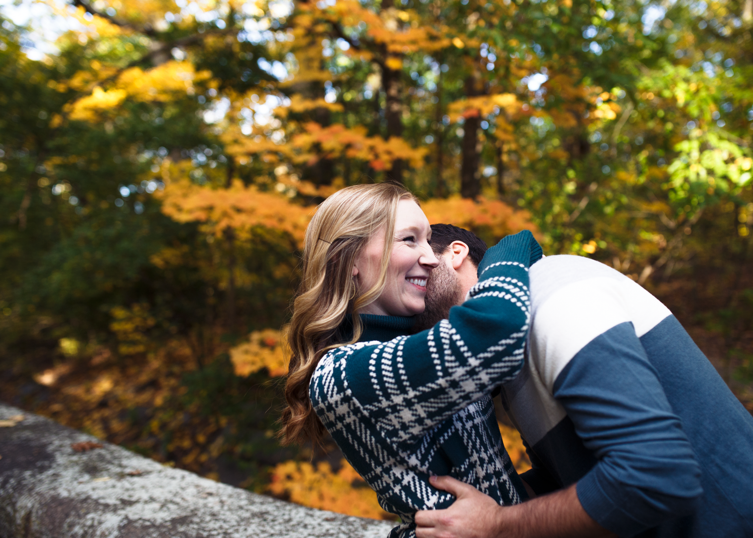 Couple embraces on a large rock surrounded by vibrant autumn foliage in a forest setting.