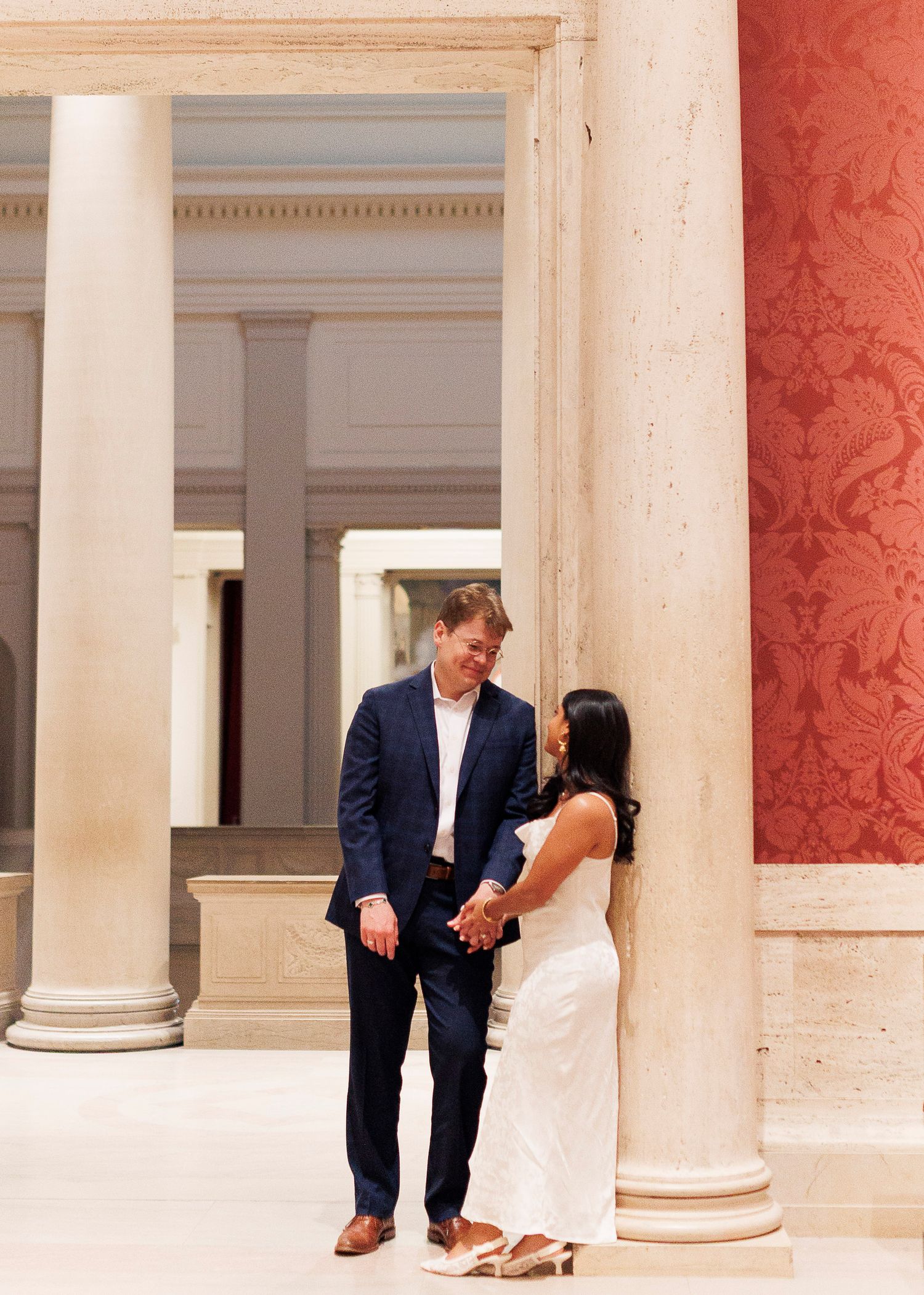 A couple in formal attire poses playfully between elegant white columns in a grand architectural setting.