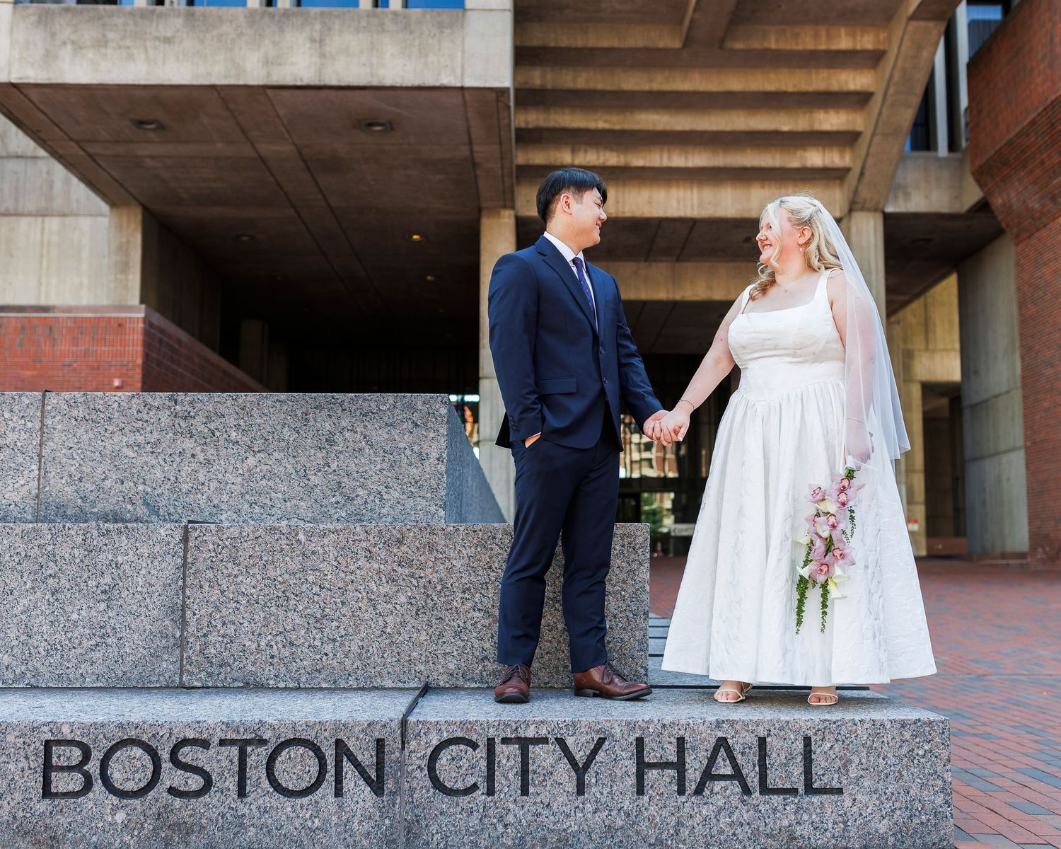 Newlywed couple holding hands at Boston City Hall, bride in white dress with veil holding bouquet, groom in navy suit.