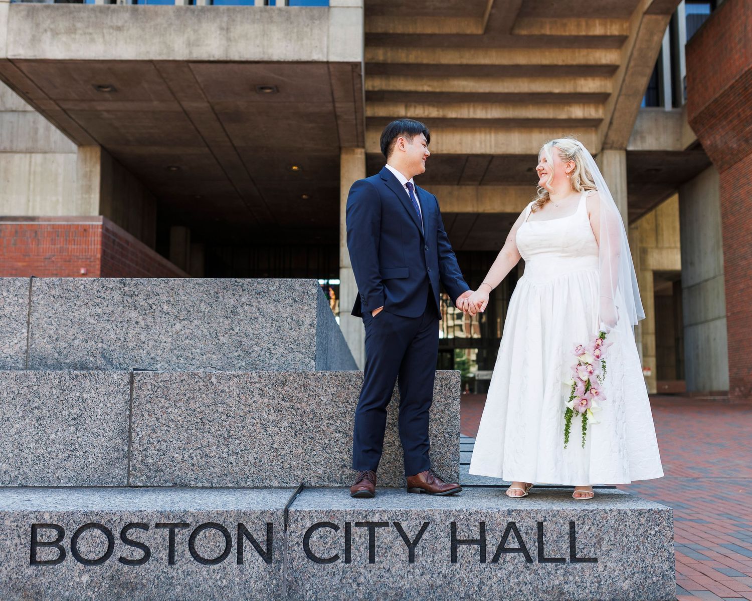 Newlywed couple holding hands on steps outside Boston City Hall, bride in white dress with veil, groom in navy suit.