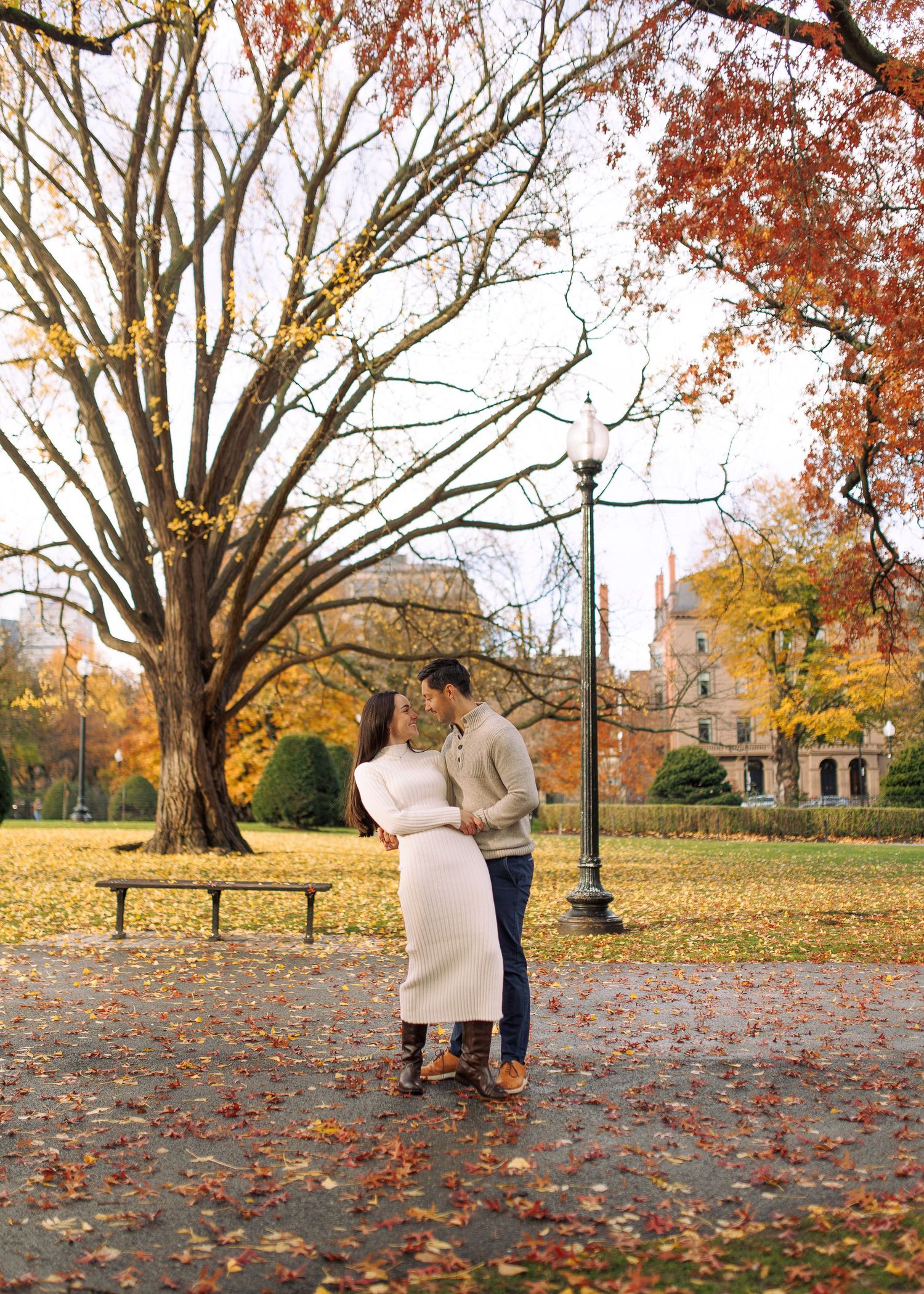 An engaged couple enjoys a romantic autumn moment under large trees with fallen leaves scattered across the ground in Boston Public Garden.