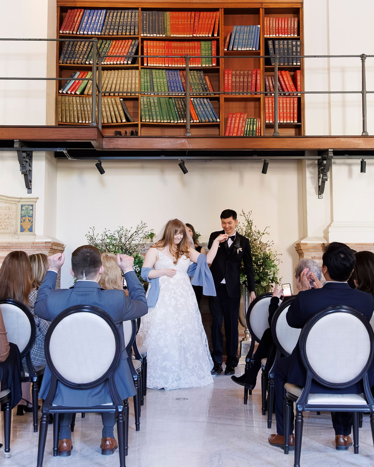 Wedding guests stand in a courtyard surrounded by orange and blue balconies during an outdoor ceremony.