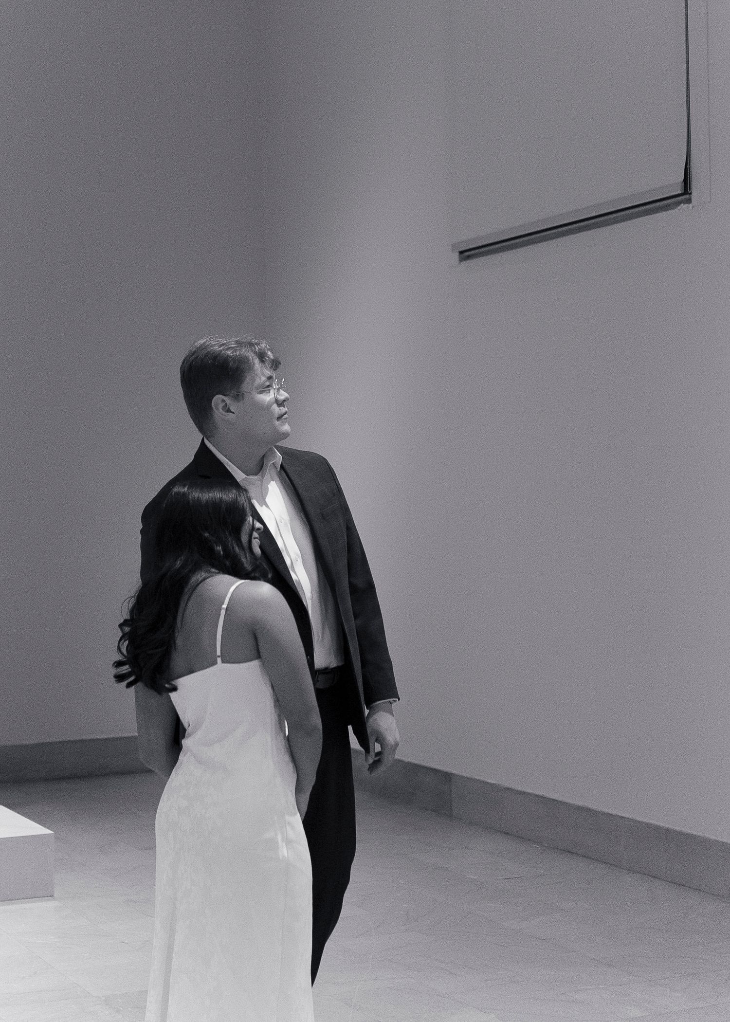 A black and white photo shows two people in formal attire standing in a minimalist hallway corner.