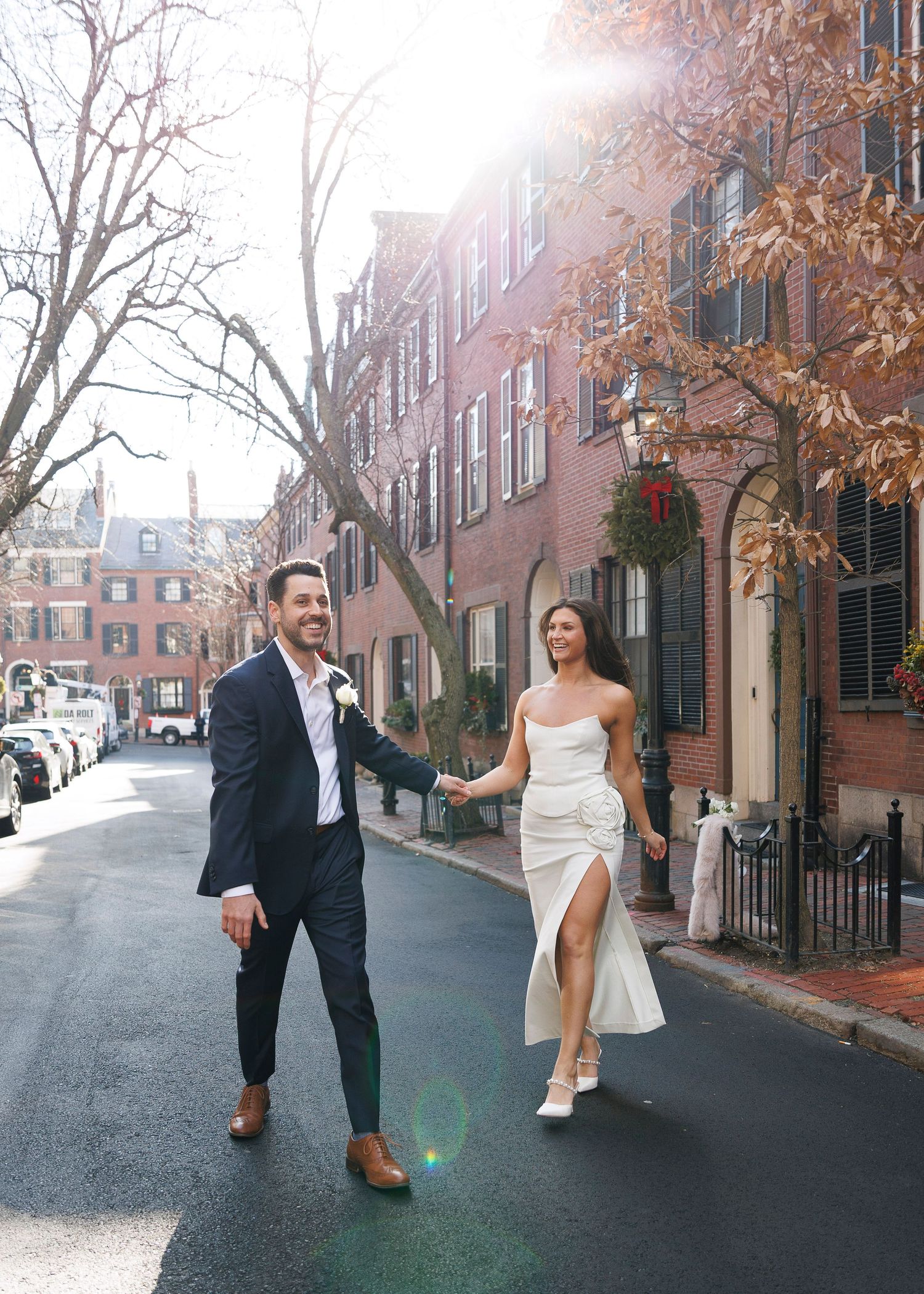 A smiling couple in wedding attire walks hand-in-hand down a sunlit brick street in Boston's Beacon Hill neighborhood.