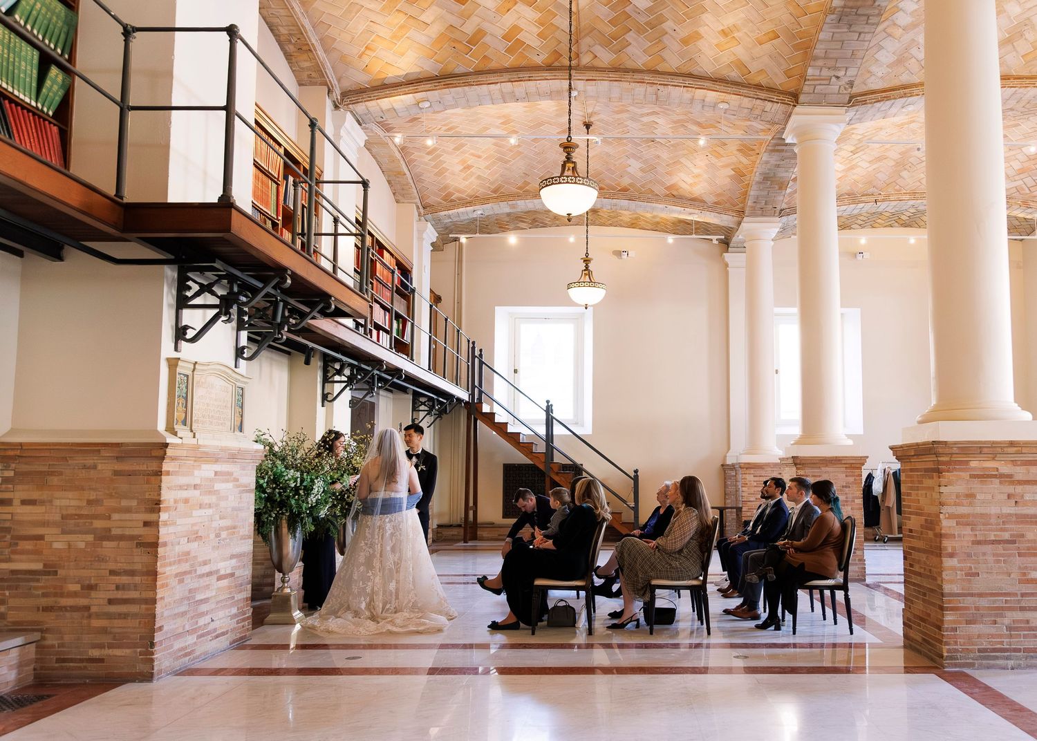 Elegant wedding ceremony in a historic venue with arched ceilings, brick columns, and guests seated in a grand hall.