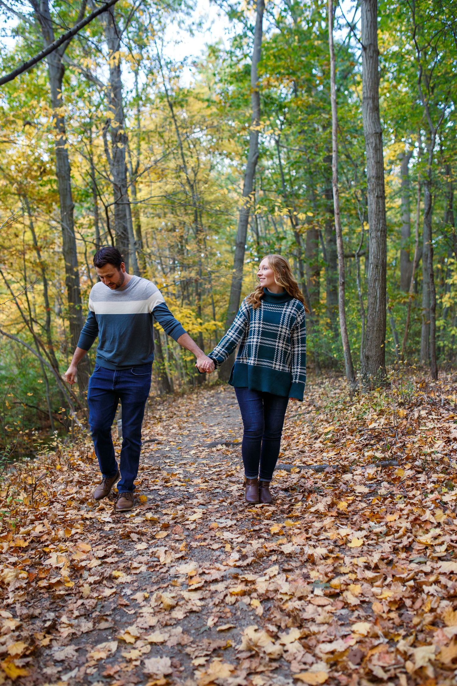 Couple holding hands walking on a leaf-covered forest path in autumn, surrounded by colorful fall foliage.