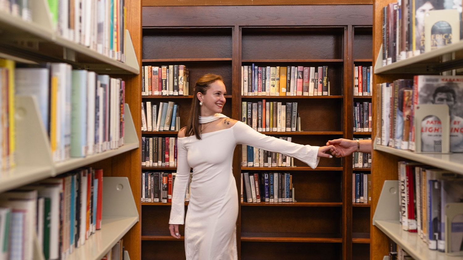 A person browses through books on wooden library shelves while standing in a long aisle lined with books.