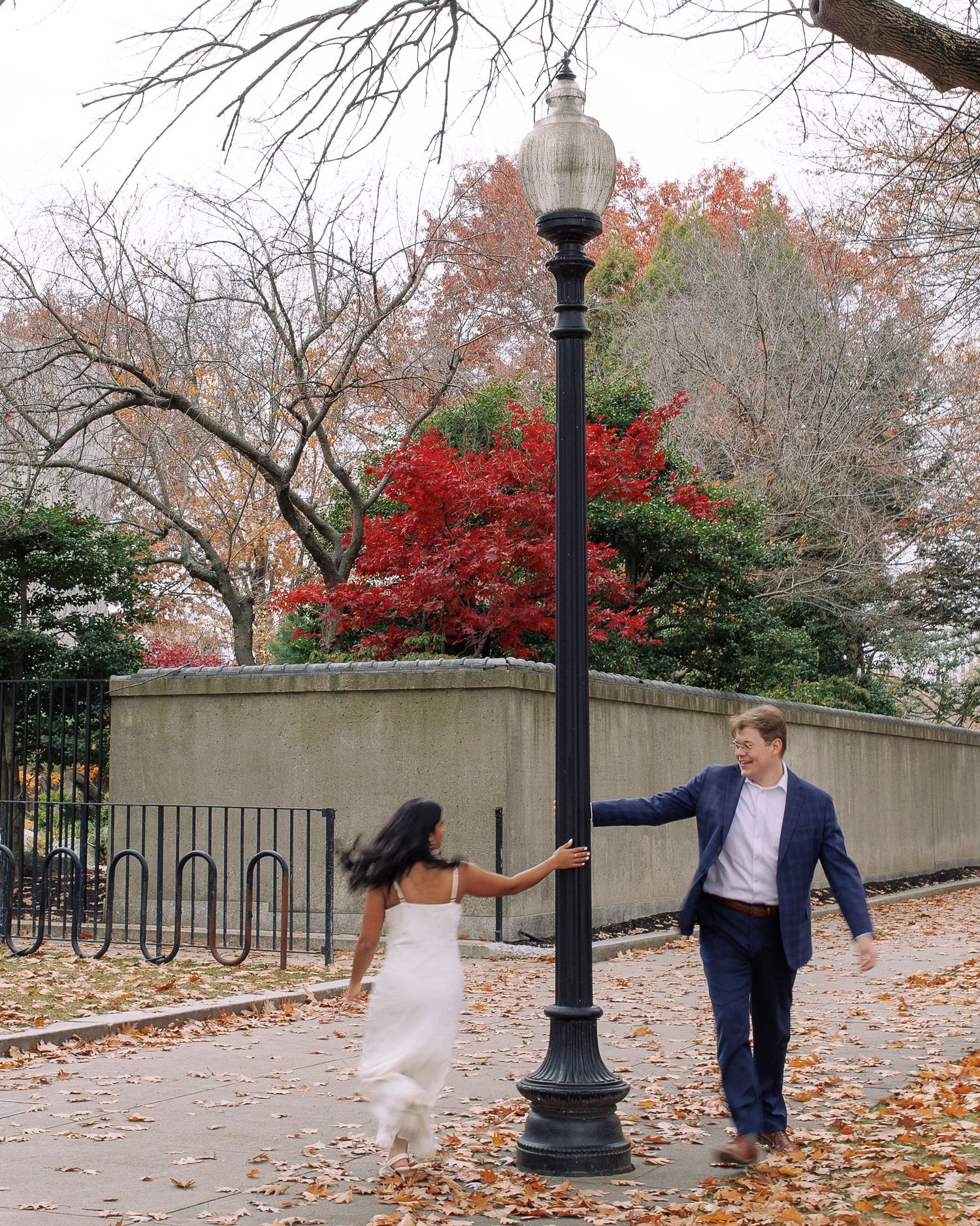 A couple dances playfully around a lamp post on an autumn day with red maple trees and fallen leaves in the background.
