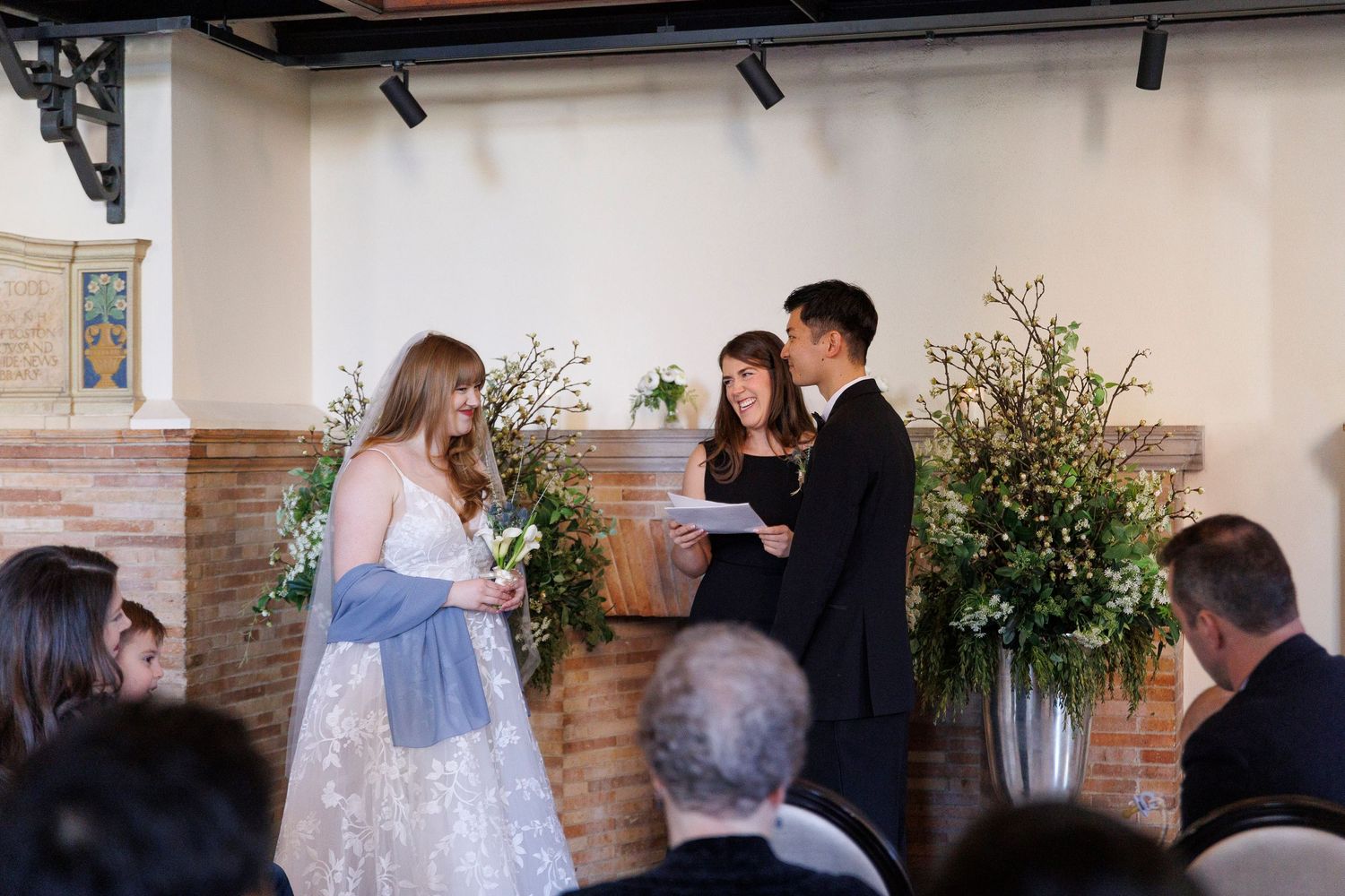 Wedding party members walk through an elegant venue hallway decorated with floral arrangements and exposed brick walls.