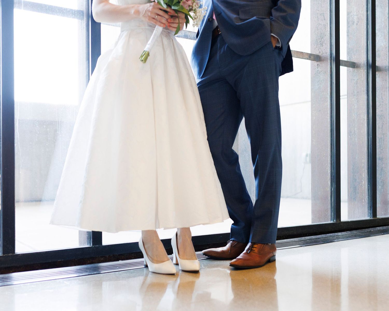 Bride in white tea-length dress and groom in navy suit standing together near large windows on wedding day.