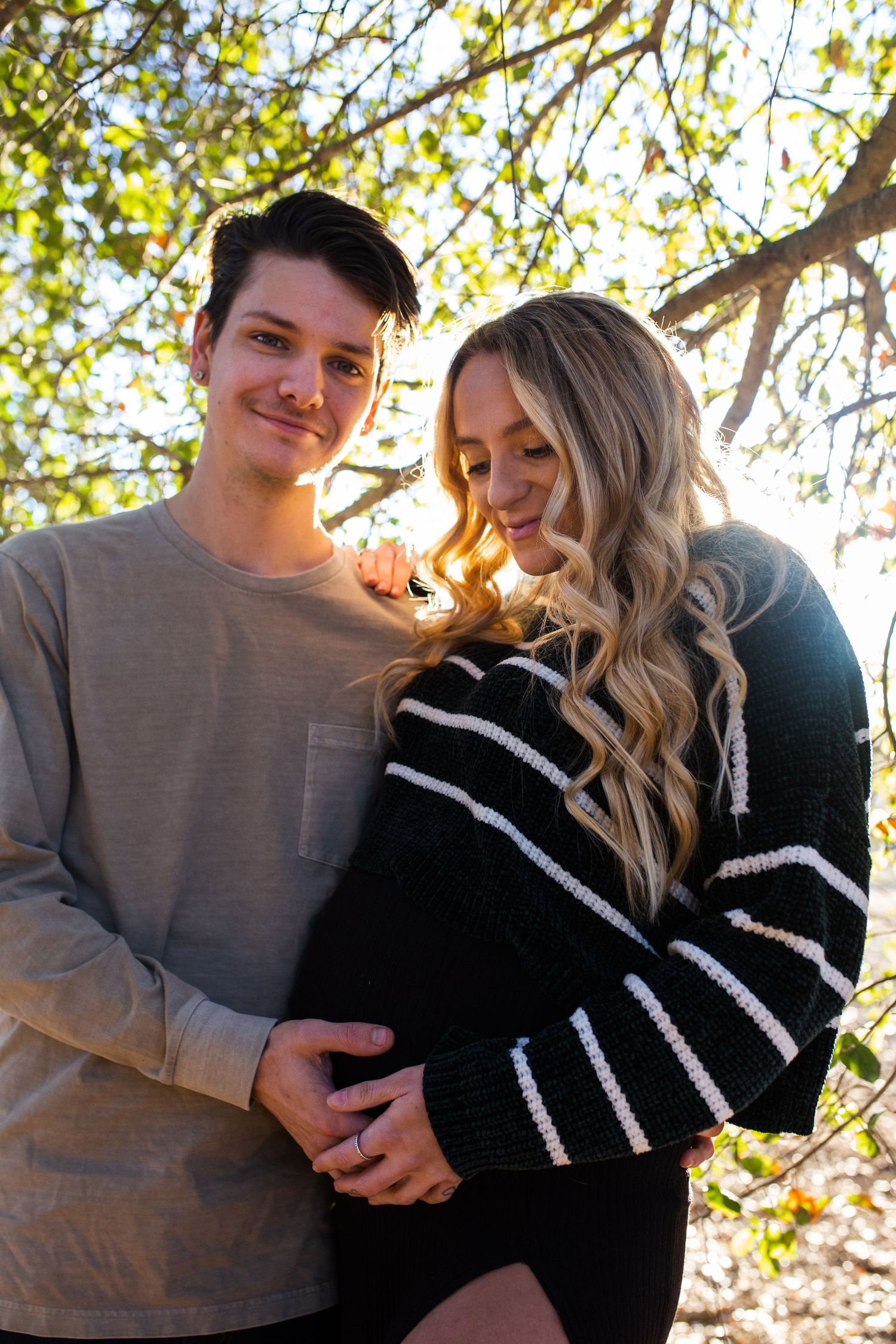 A couple embraces outdoors under autumn tree branches with golden sunlight filtering through the leaves.