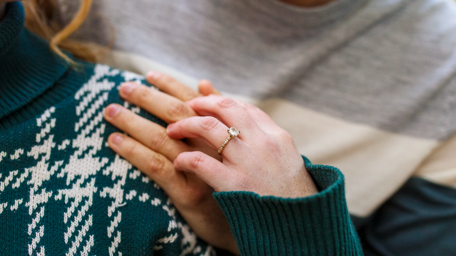 Hands wearing a green and white houndstooth sweater with a delicate engagement ring on display.