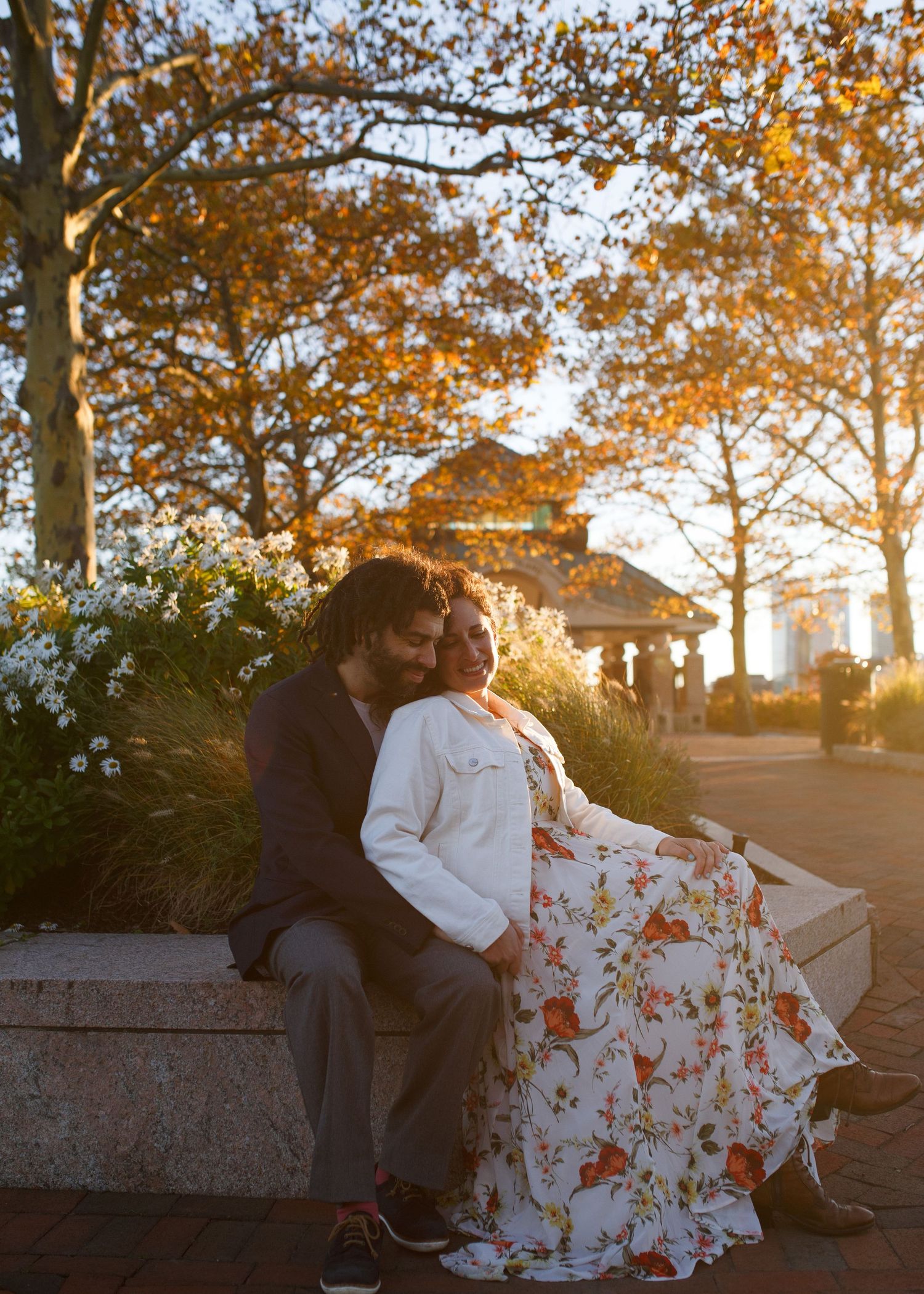 A couple shares a romantic moment on a stone wall at sunset while autumn leaves create a warm golden backdrop.