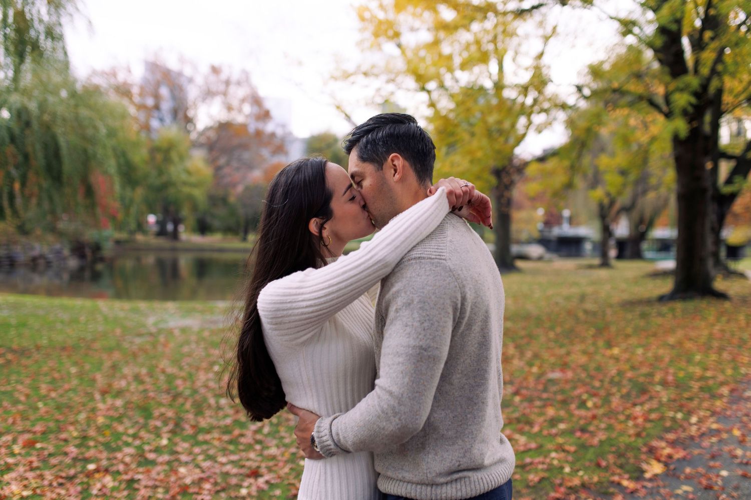 Romantic couple in cozy sweaters embracing outdoors during autumn with colorful fall foliage in the background at Boston Public Garden.