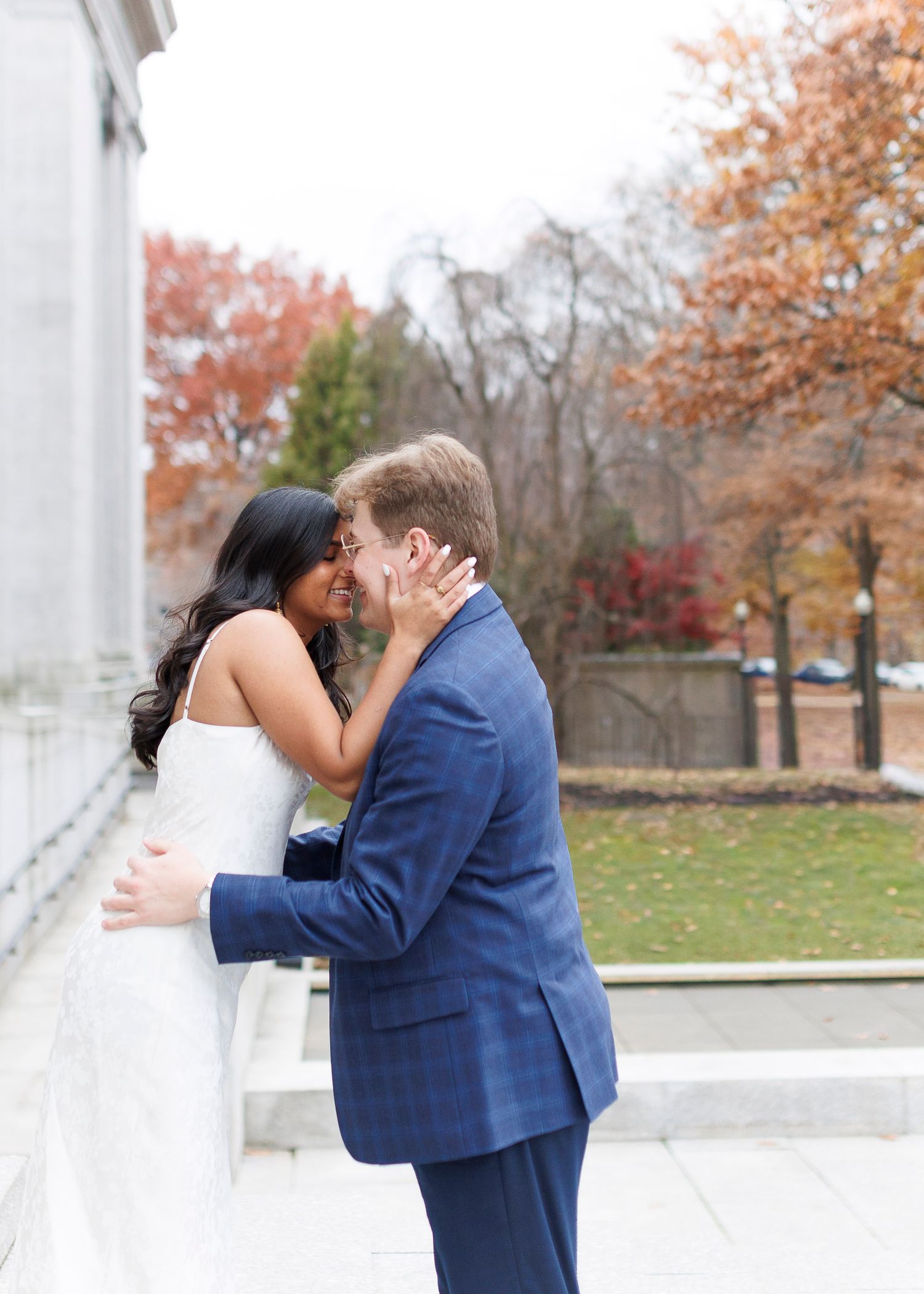 A couple shares a romantic kiss in front of white columns during an autumn day with colorful fall foliage in the background.