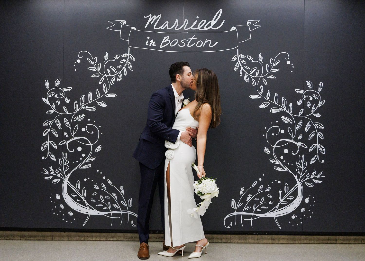 Newlywed couple kissing in front of a chalkboard sign reading Married in Boston with floral designs.