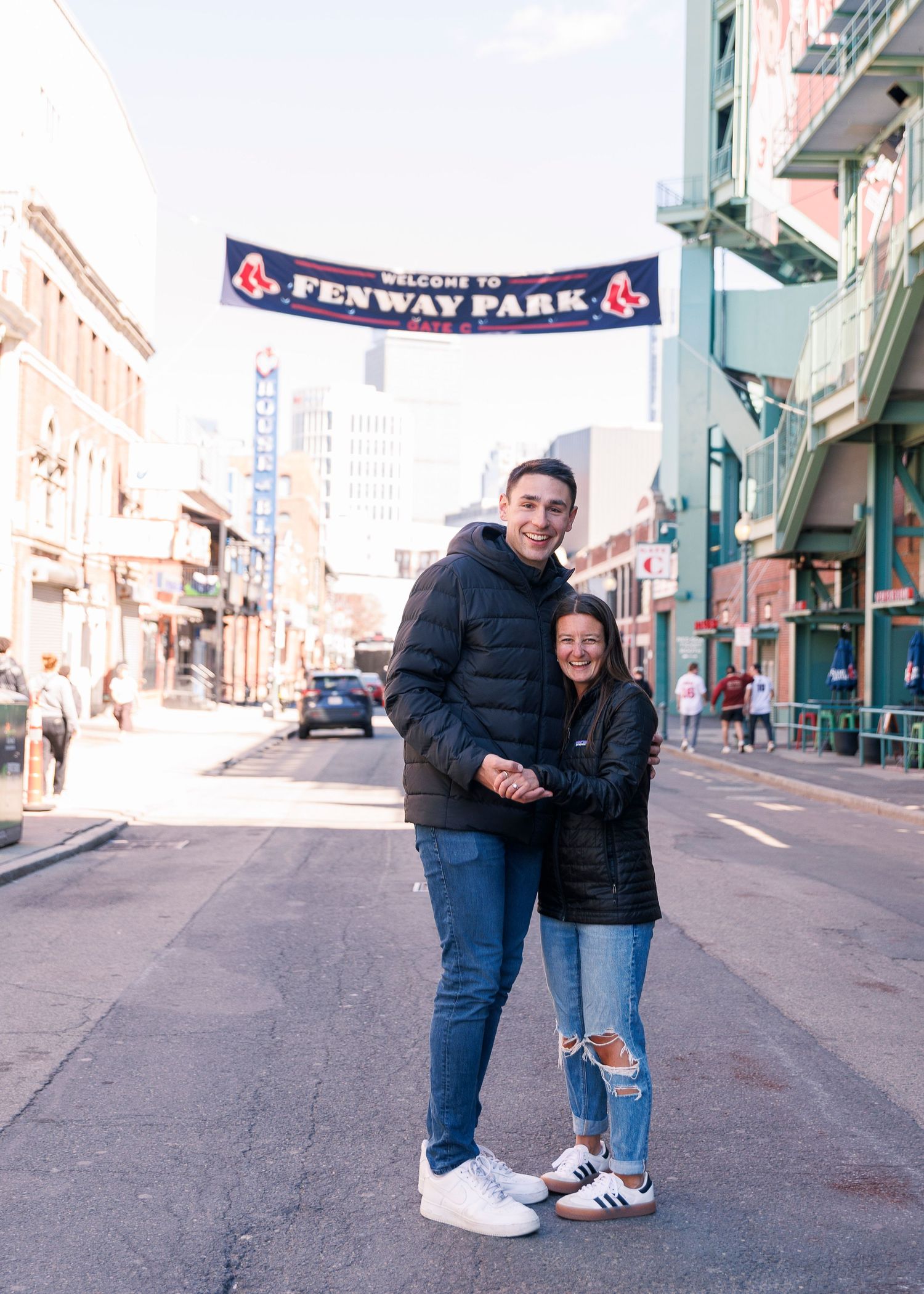 Couple smiling and posing together on a street outside Fenway Park in Boston, wearing black jackets and jeans.