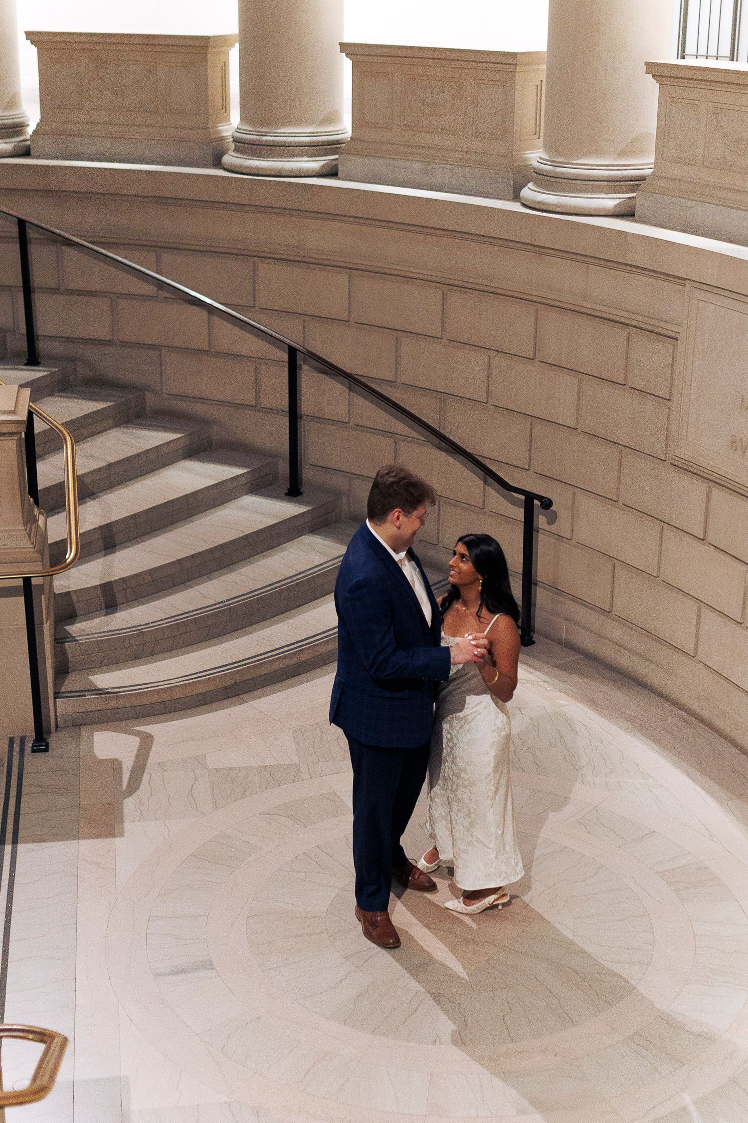 Couple in formal wedding attire sharing intimate moments on curved marble staircase in elegant venue.