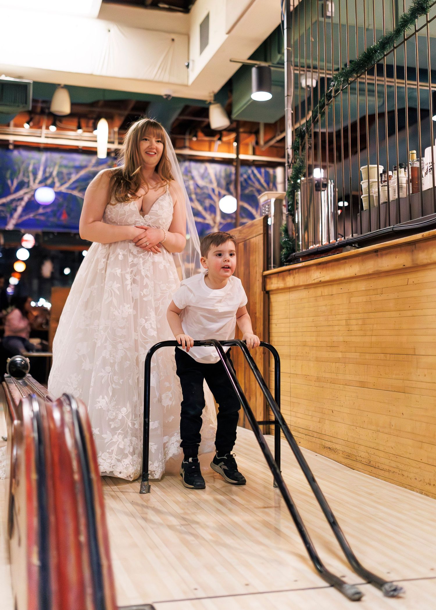 A parent and child stand together at a bowling alley lane, with decorative lights and railings visible in the background.