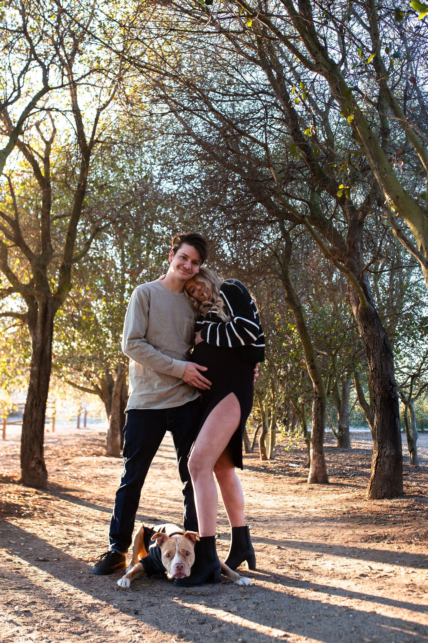 A couple poses with their small dog on a tree-lined path during golden hour sunset in a park.